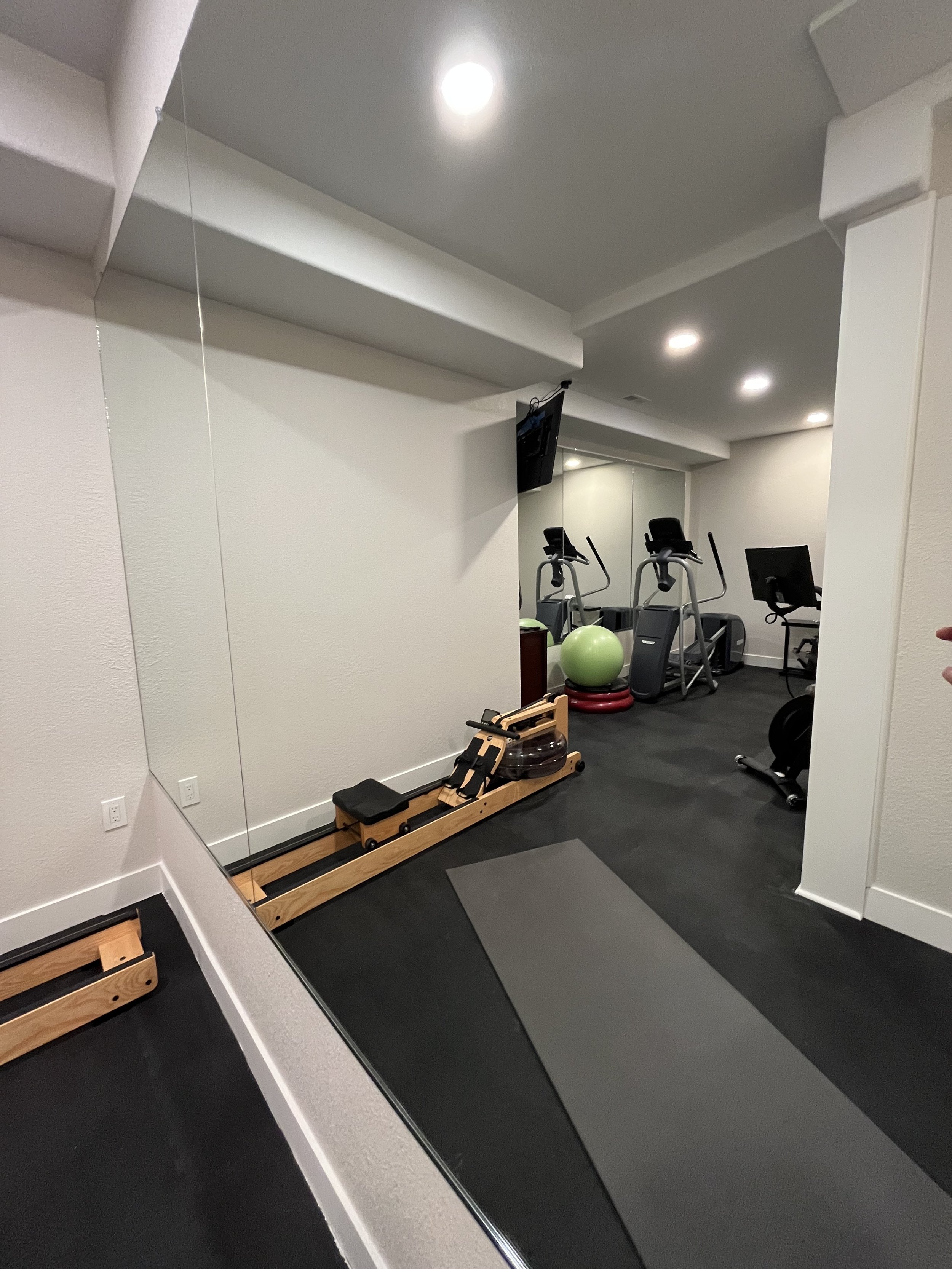 Photo of a small home gym with exercise equipment including elliptical machines, a chair, a mirror, a green exercise ball, and a yoga mat on a dark floor. Recessed ceiling lights illuminate the space.