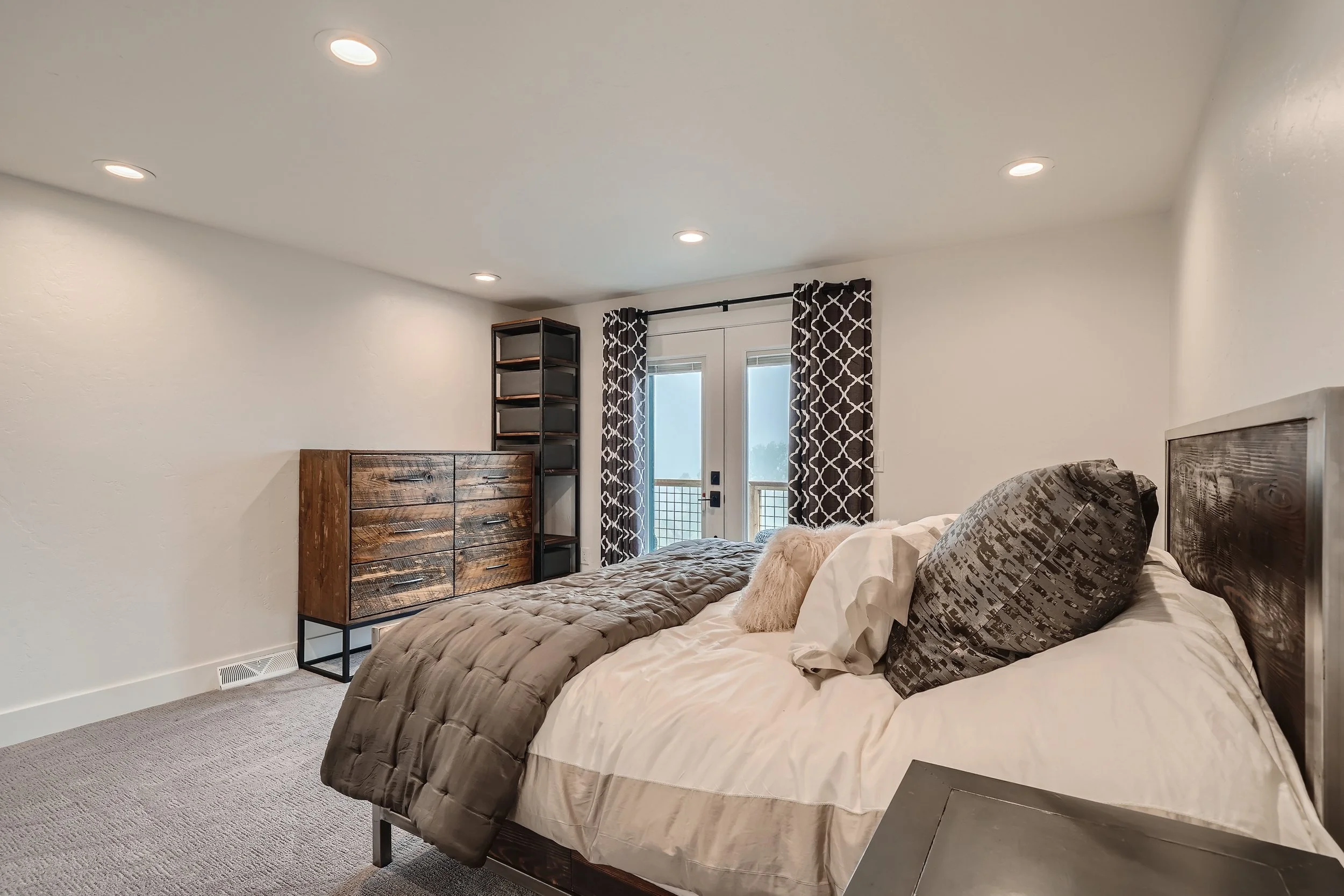 A bedroom with a bed, pillows, and a wooden dresser, with a balcony door and patterned curtains in the background.