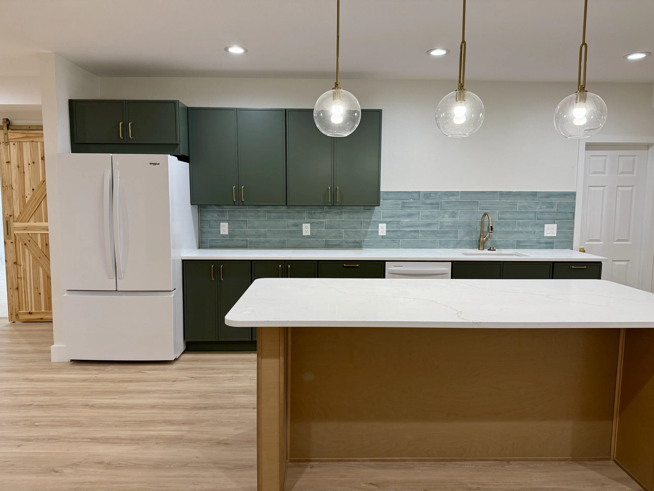 Modern kitchen with green cabinets, white marble island, blue subway tile backsplash, stainless steel faucet, and white appliances, illuminated by three pendant lights.
