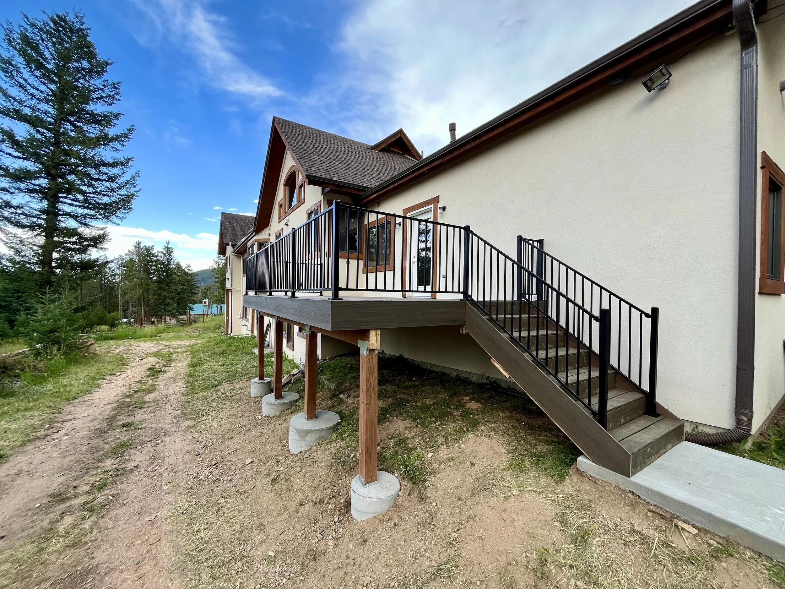 Exterior view of a house with a wooden staircase and balcony, surrounded by trees and a dirt road.
