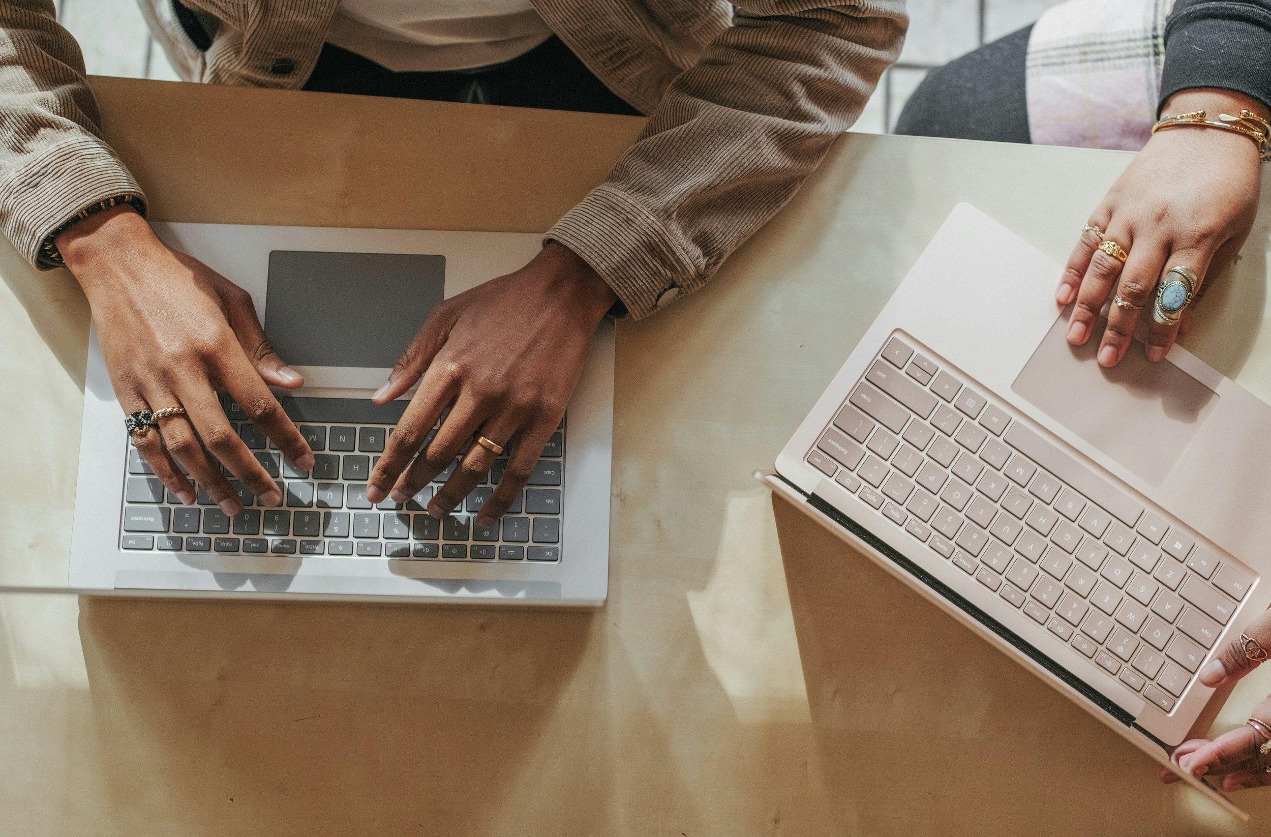 Two people sitting at a light wood table using laptops. One person's hands are on a silver keyboard laptop, and another person's hands are on a pink keyboard laptop. Both individuals are wearing jewelry and have rings on their fingers.