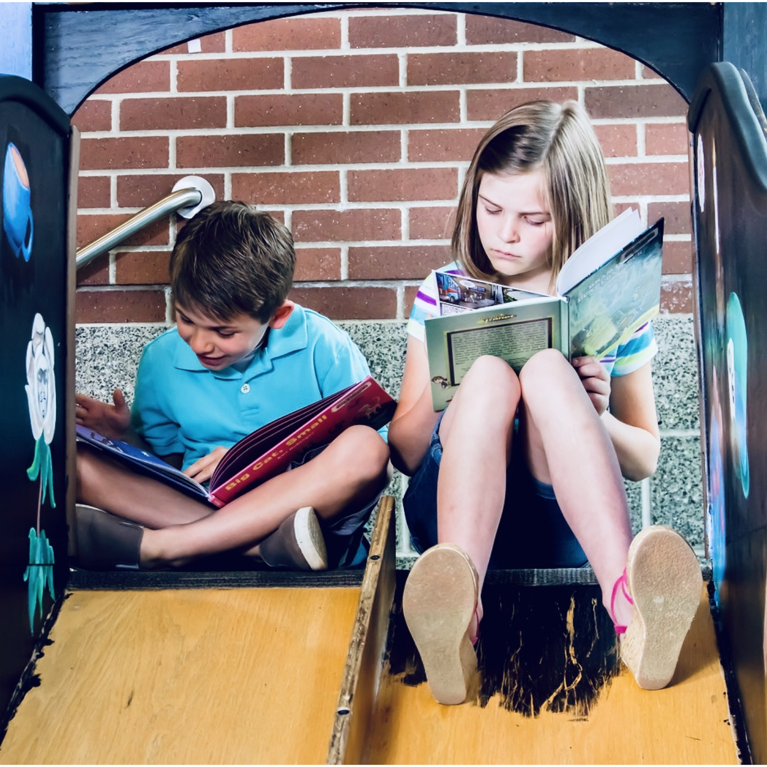 Two children, a boy and a girl, sitting on a wooden slide reading books, with a brick wall in the background.