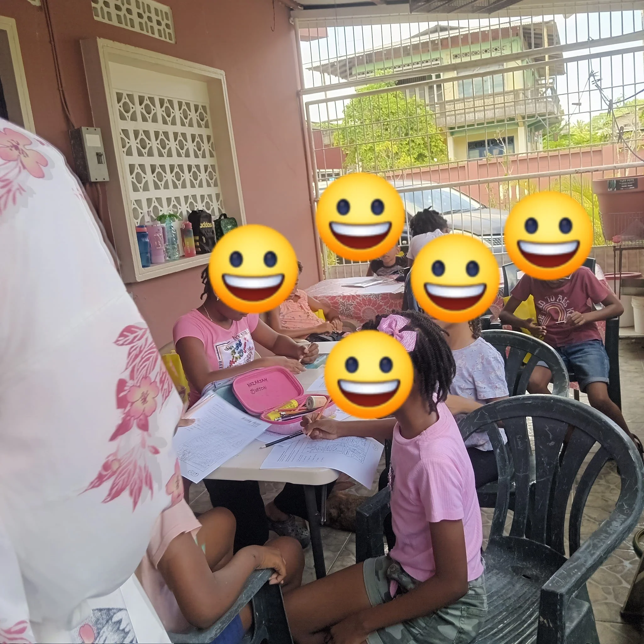 Children sitting at a table on an outdoor patio, engaging in activities or work, with a pink wall, a decorative white lattice, and a metal fence in the background.