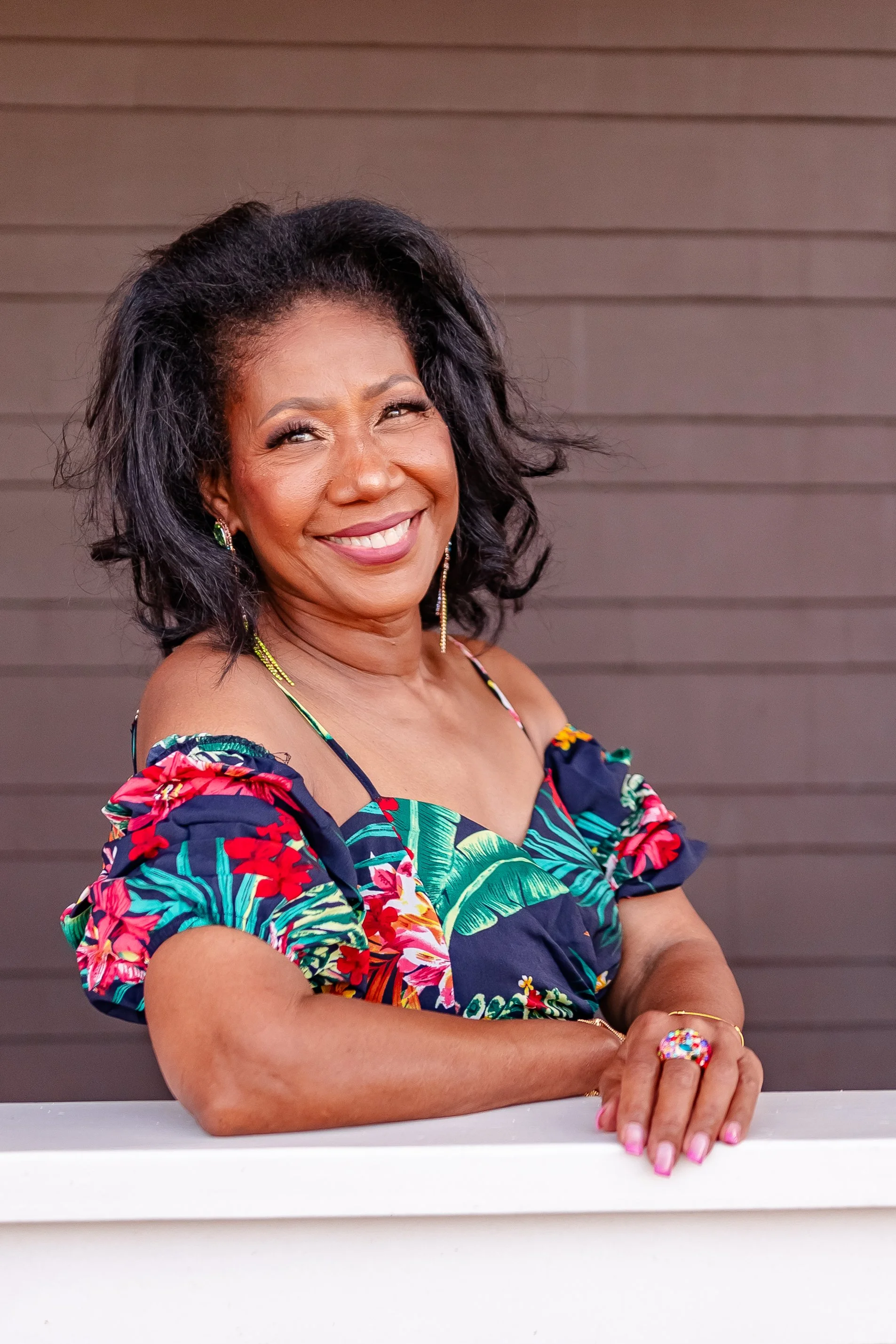 Smiling woman with dark hair, wearing a colorful tropical dress with floral and leaf patterns, standing in front of a gray horizontal siding wall.