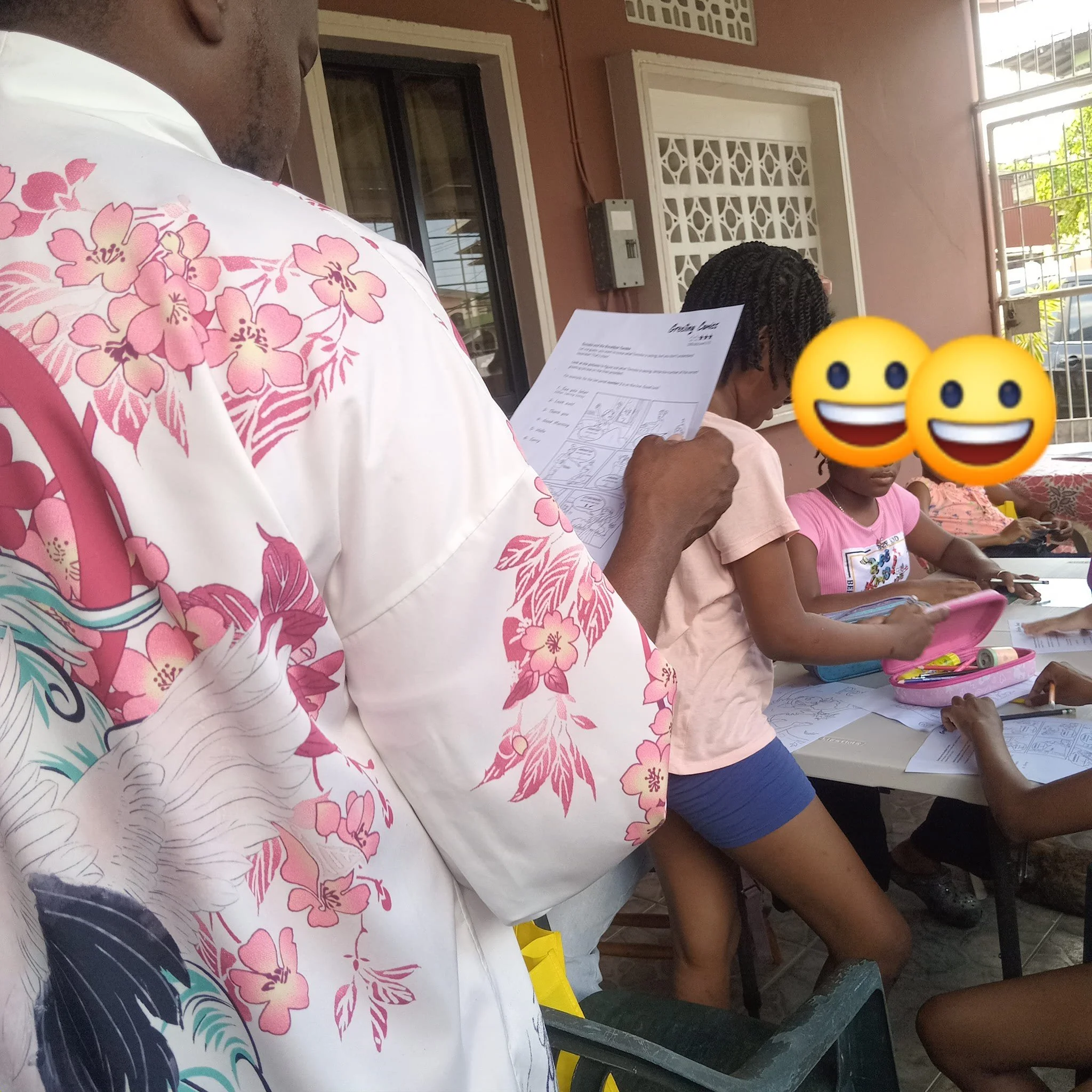 A group of children sitting at a table with books and papers, with an adult standing nearby holding a sheet of paper. The children's faces are covered with smiley face emojis, and they appear to be engaged in an educational activity.