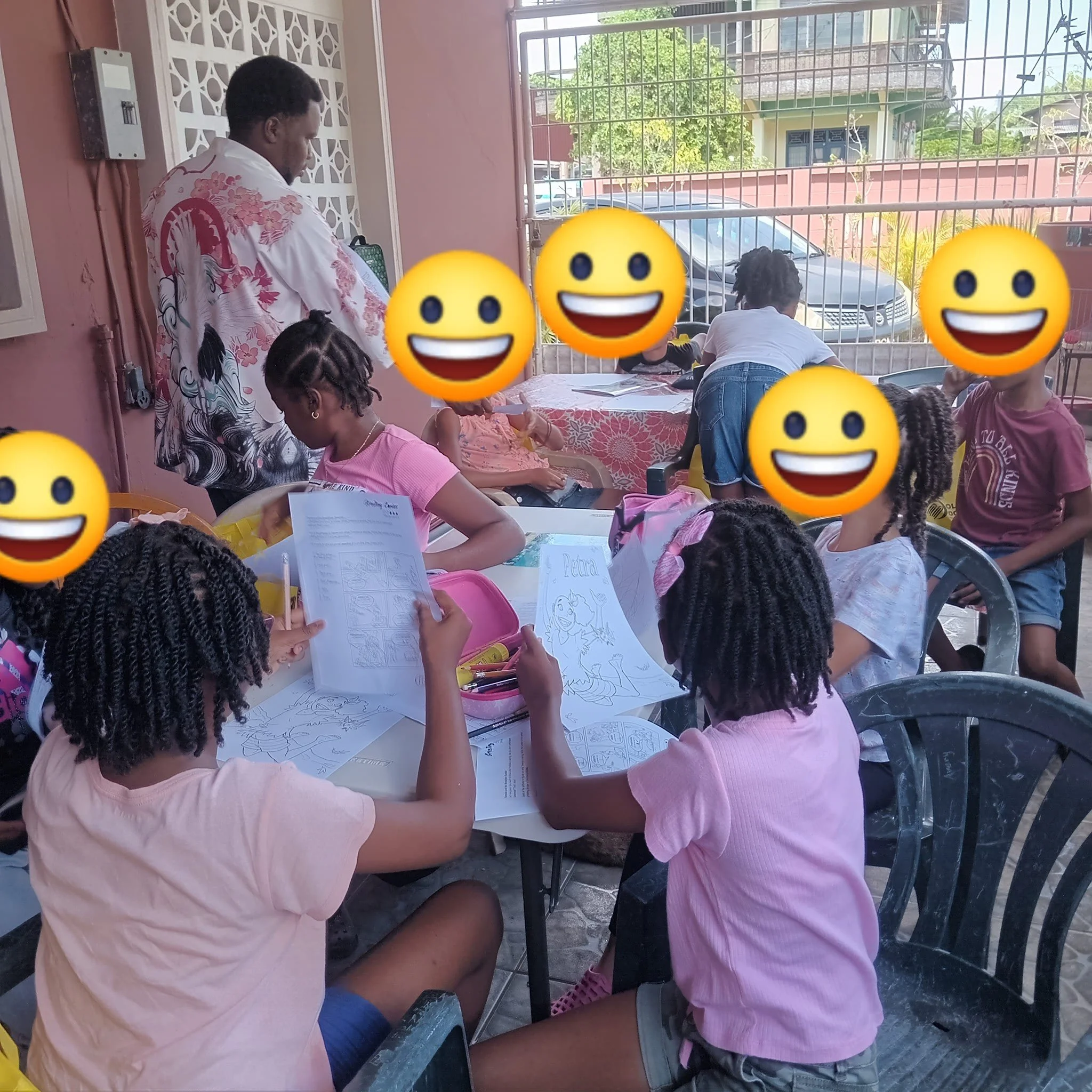 Children sitting at plastic tables outside, coloring and looking at coloring books, with a woman supervising, in a residential area with a fence and houses in the background.