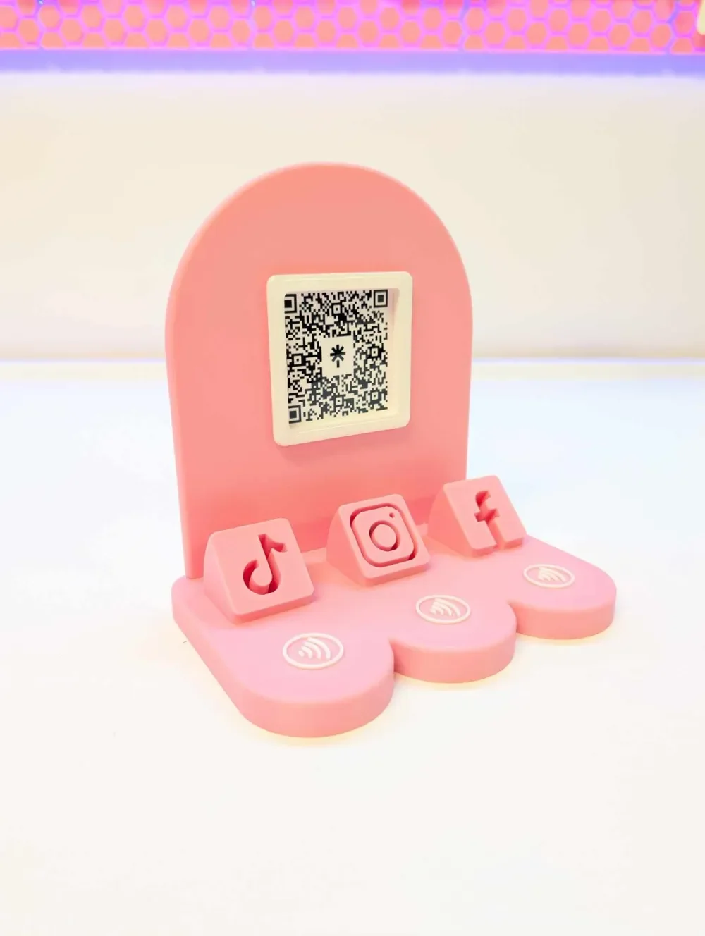 Pink phone-shaped stand with QR code on the screen and social media icons for TikTok, Instagram, and Facebook on the base.