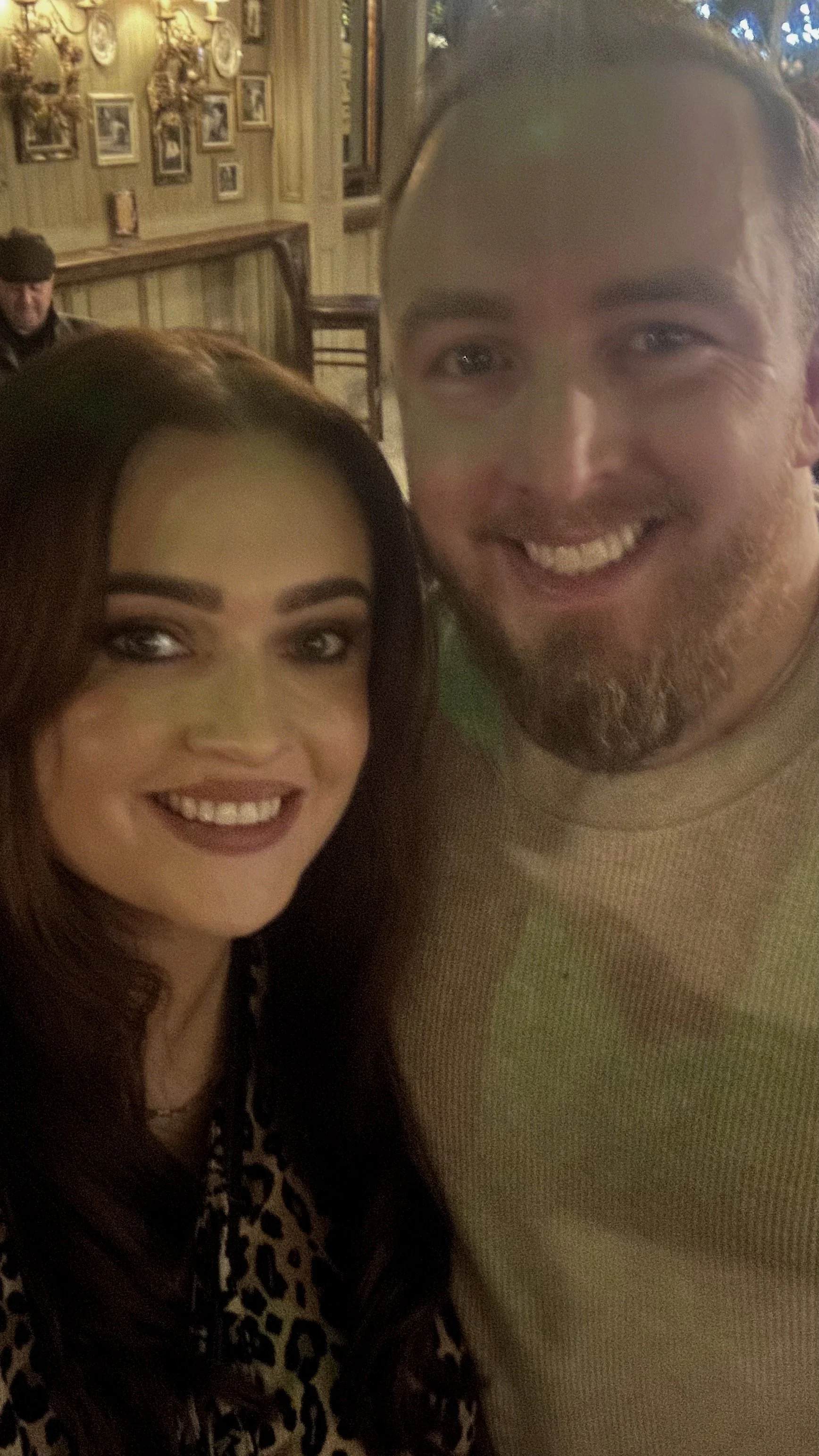 A woman with long brown hair and a man with a beard smiling and taking a selfie in a warmly lit indoor setting with framed pictures on the wall.