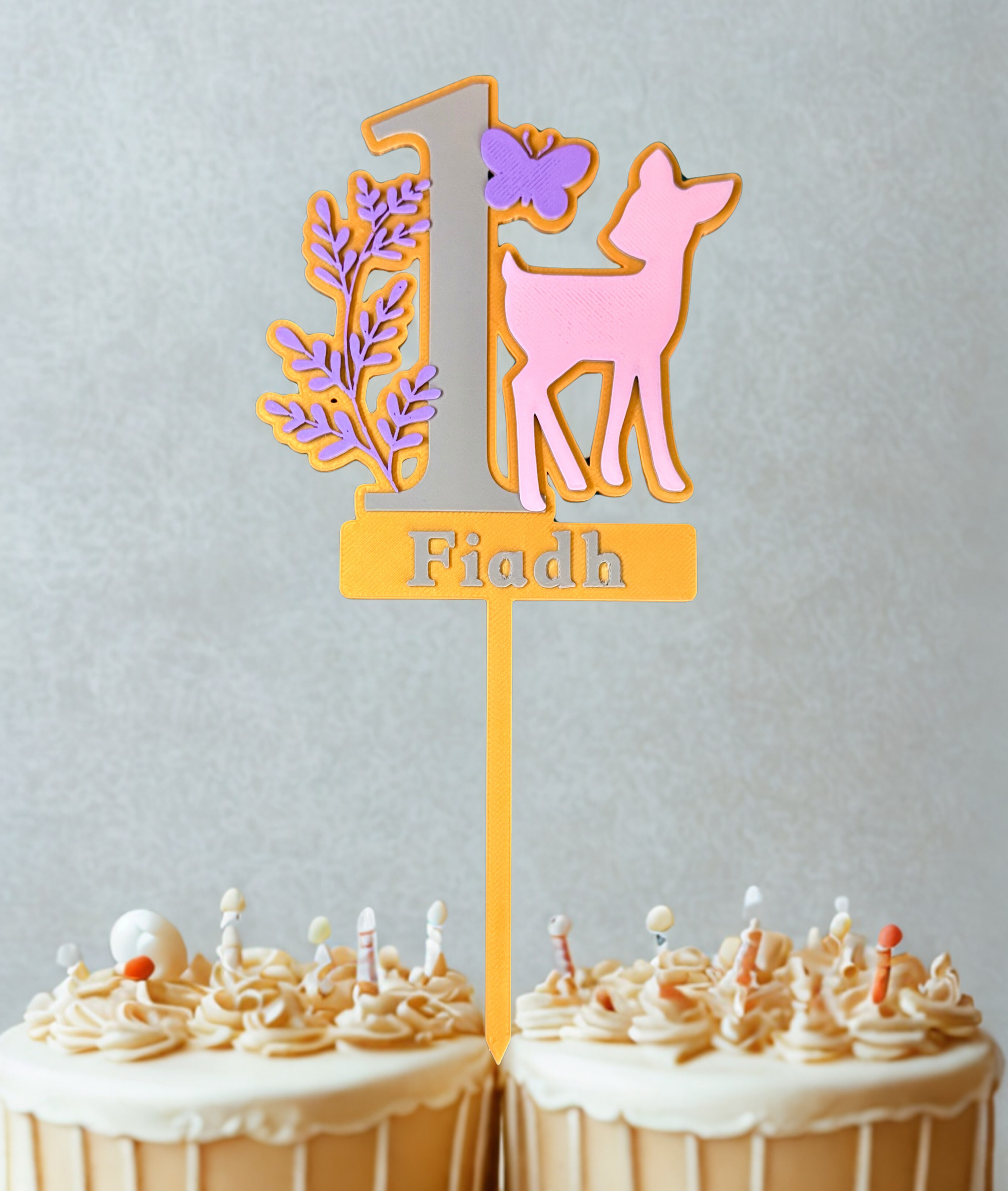Two cupcakes with beige frosting and sprinkles, topped with a decorative topper that has the number 1, a butterfly, leaves, and a pink animal silhouette, with the name "Fiadh" on a yellow banner.