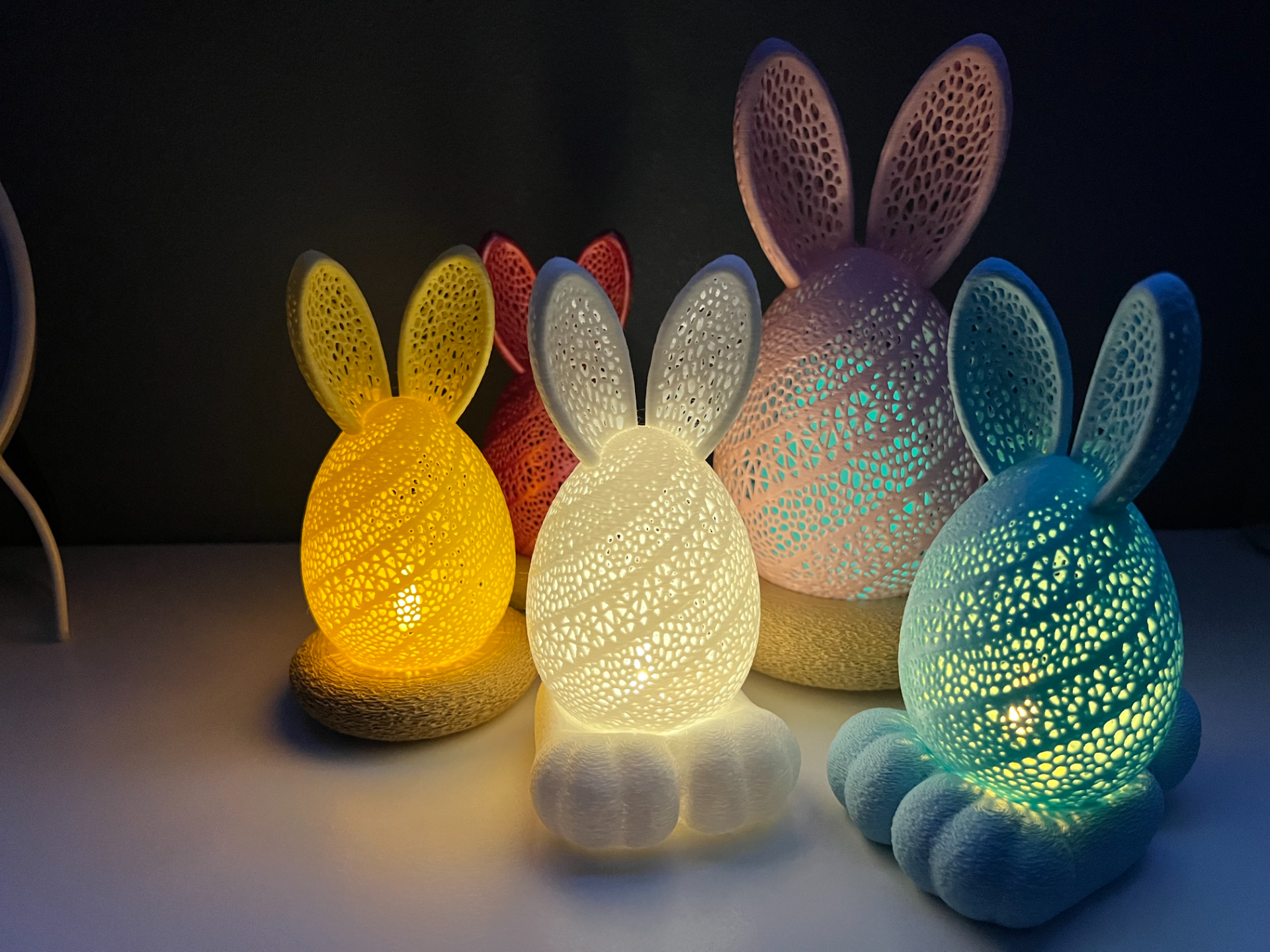 Colorful illuminated bunny and egg-shaped decorations with bunny ears, made of intricate lattice designs, glowing in yellow, white, pink, blue, and purple, arranged on a flat surface against a dark background.
