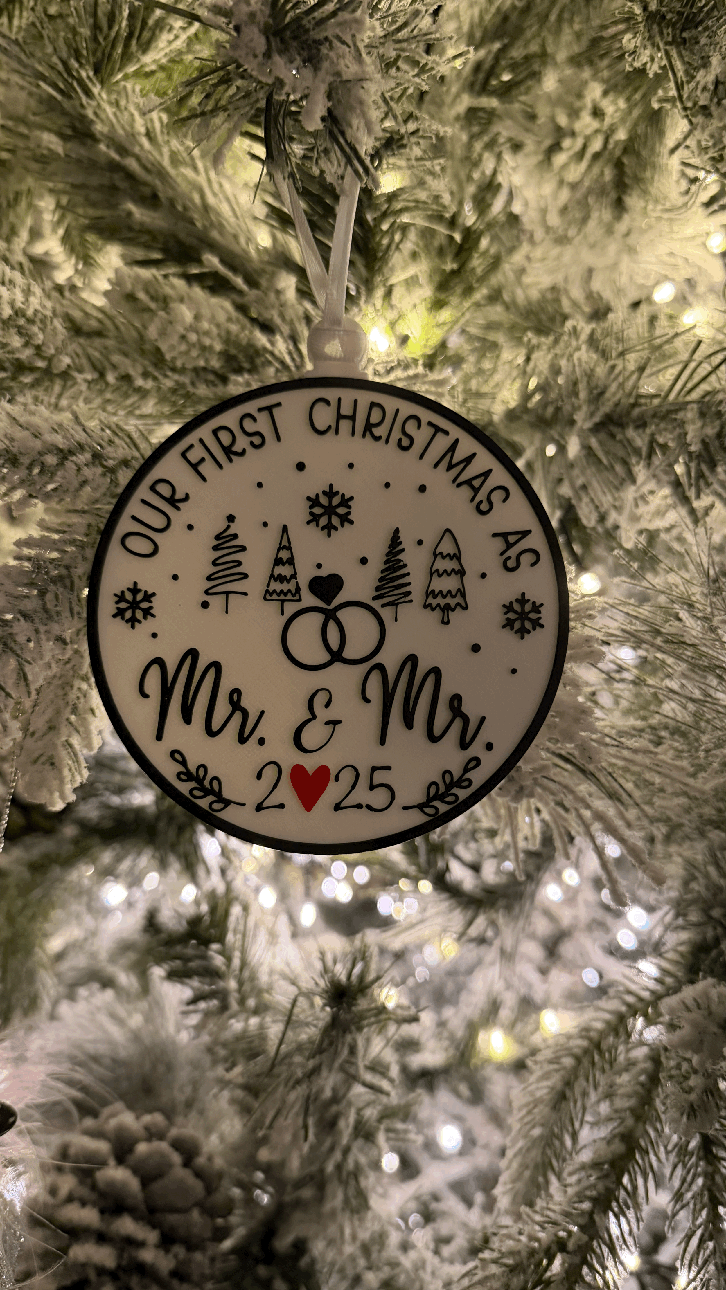 A Christmas tree ornament with the inscription 'Our First Christmas As Mr. & Mrs. 2025,' decorated with snowflakes, trees, and wedding rings, hanging on a snow-flocked Christmas tree.