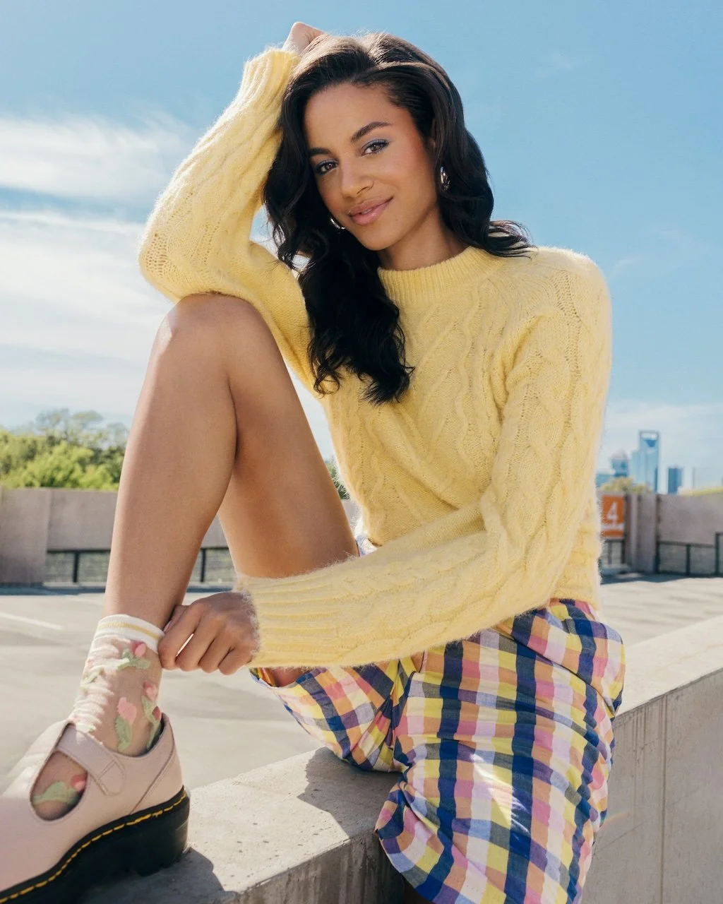 A young woman sitting outdoors on a ledge, wearing a yellow sweater, plaid shorts, floral socks, and chunky shoes, with her hair styled in loose waves and a confident smile.