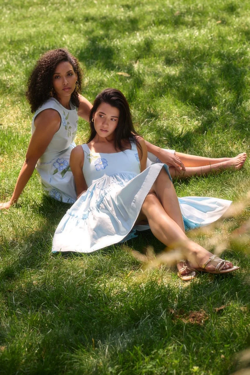 Two women in light blue dresses with floral patterns sitting on grass in a park, surrounded by shaded trees.