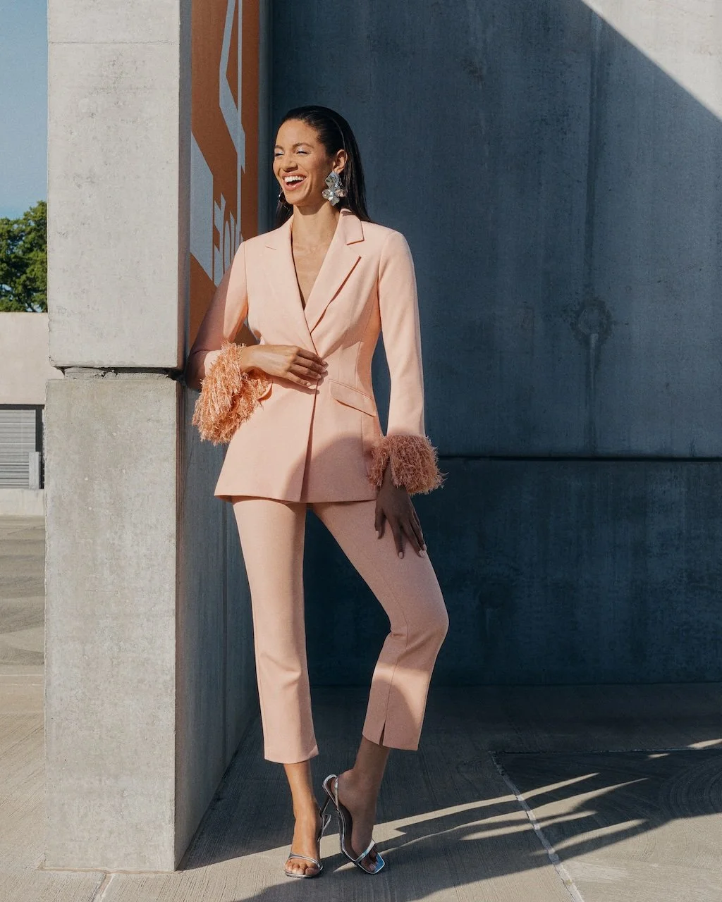 A woman in a peach-colored suit with feathered cuff details, standing outdoors against a concrete wall, smiling, wearing high heels and large earrings.