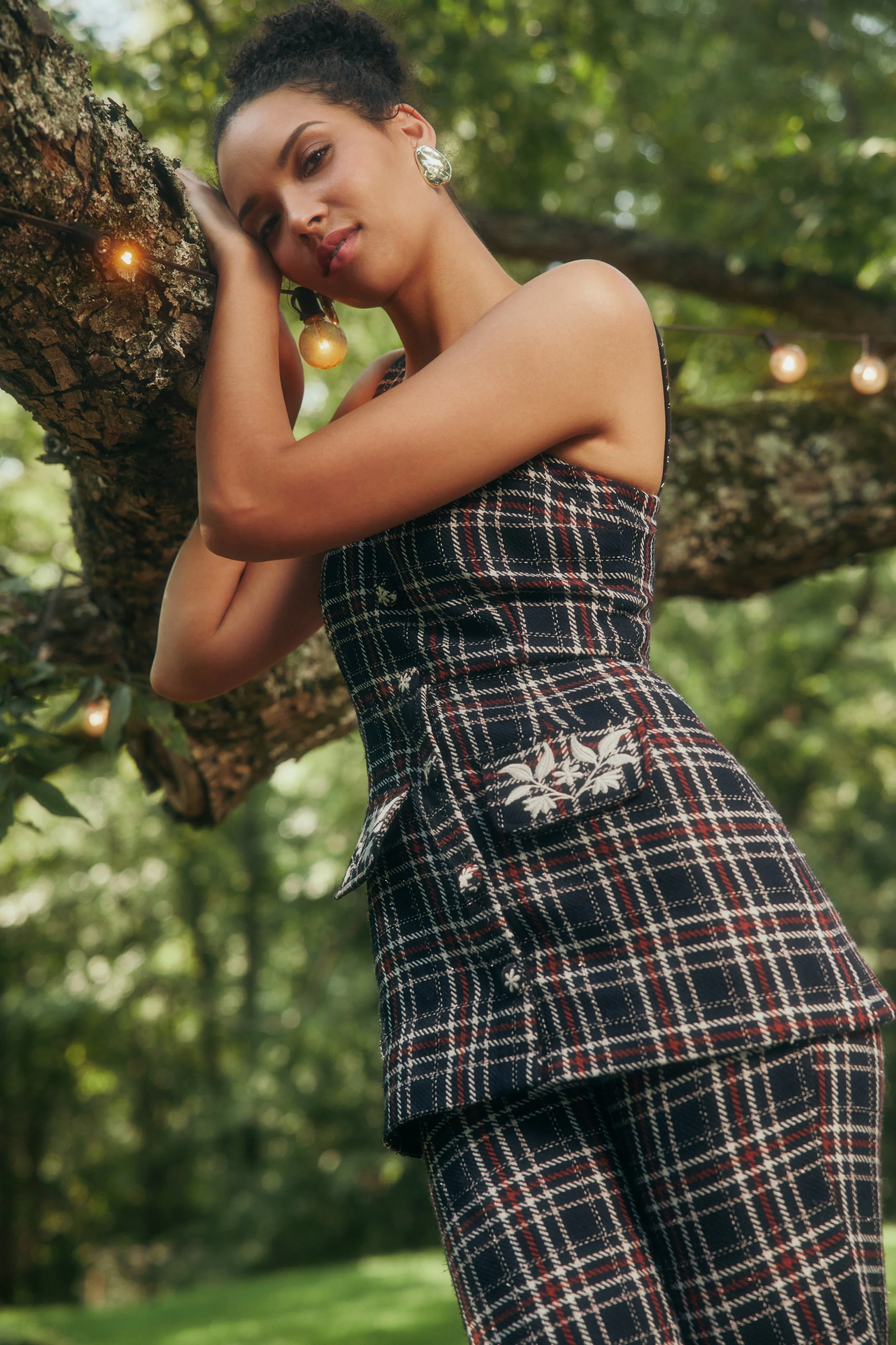 A woman with dark hair tied back, wearing earrings and a plaid outfit with floral embroidery, leans against a tree in a green outdoor setting with string lights.