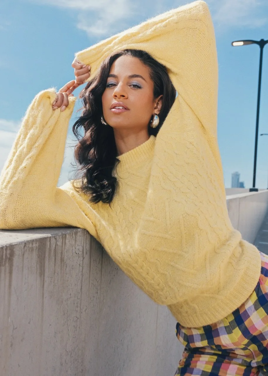 A woman with dark, wavy hair, wearing a yellow sweater and colorful plaid pants, posing outdoors during daytime with a city skyline in the background.