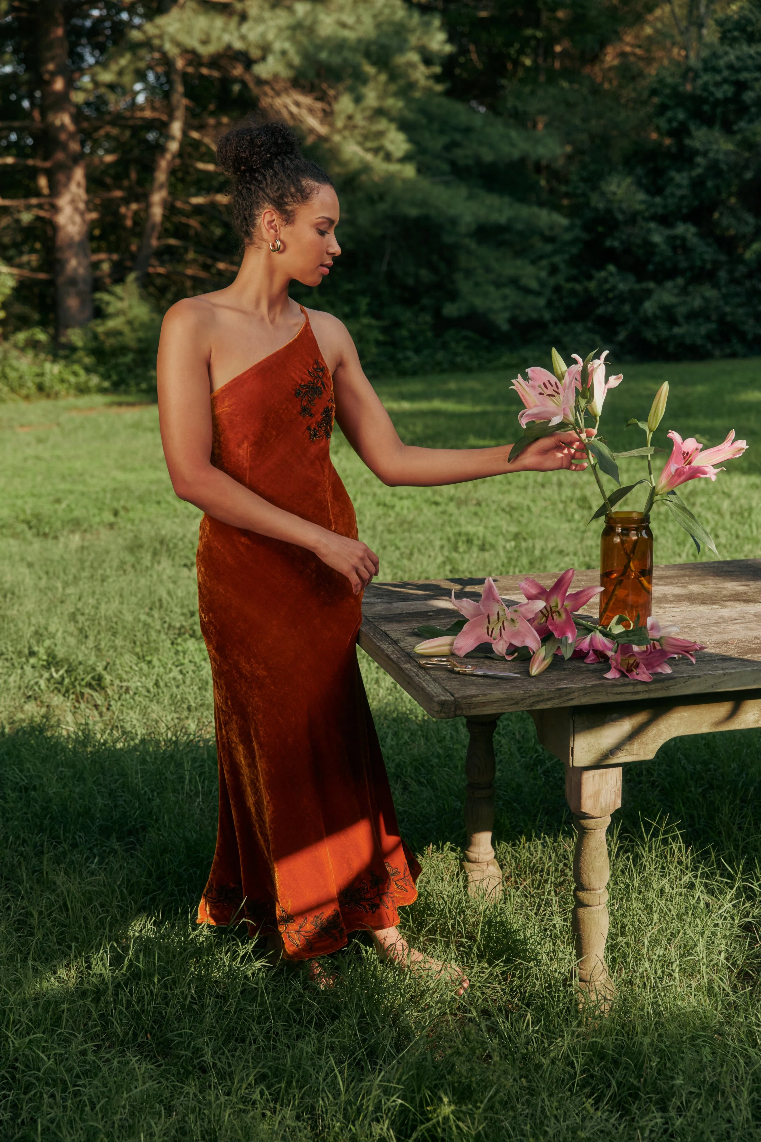 A woman in a burnt orange velvet dress arranging pink lilies in a brown glass jar outdoors in a grassy area with trees in the background.