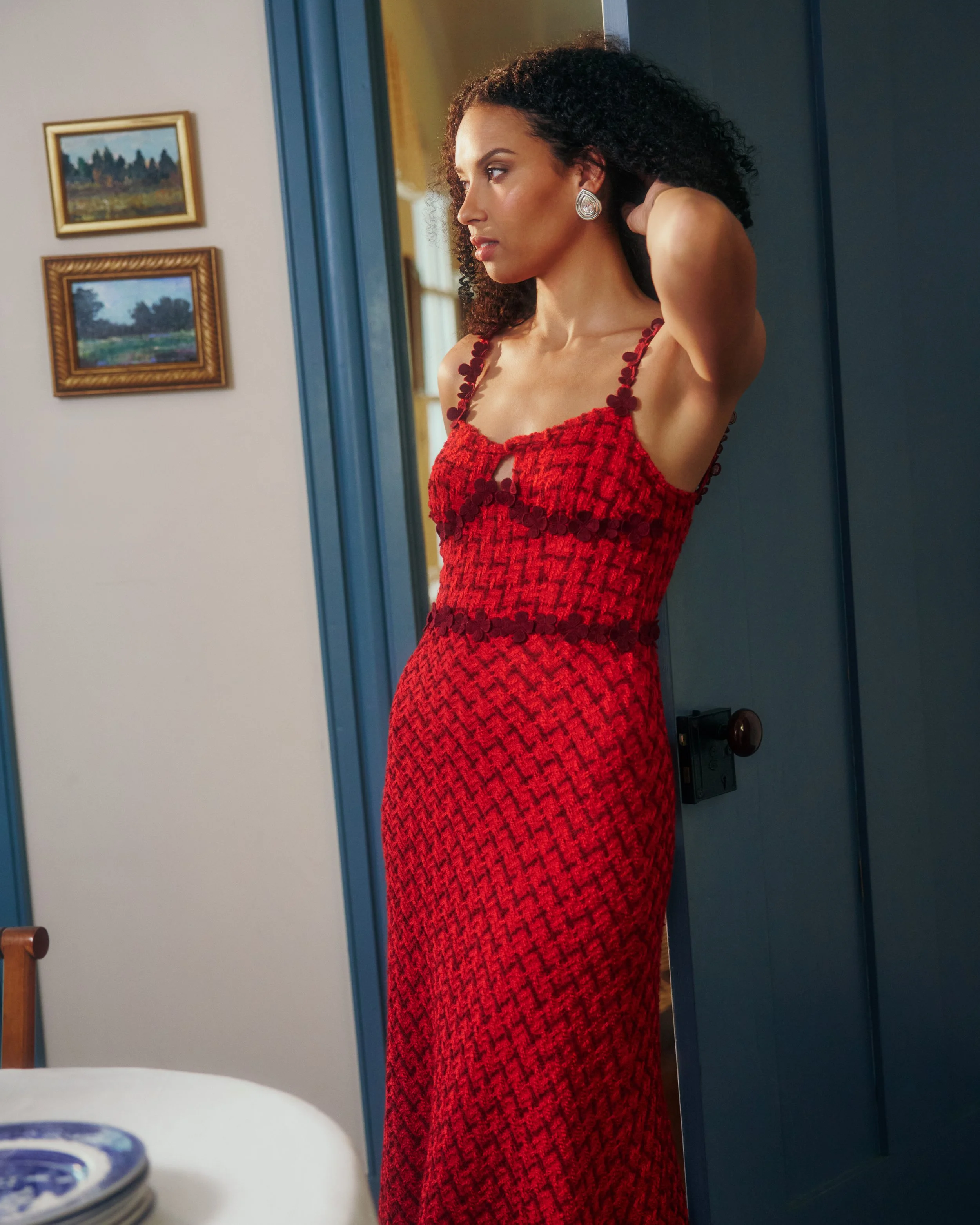 A woman with curly hair in a red patterned dress standing near a blue door, adjusting her hair, in a room with framed landscape paintings on the wall.