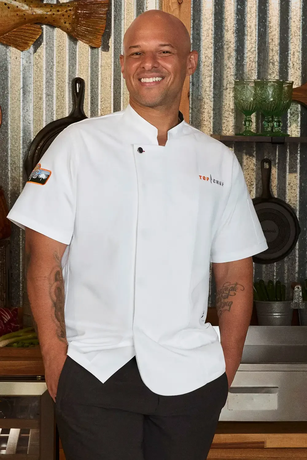 A smiling chef with tattoos on his right arm, wearing a white chef coat with a logo and black pants, standing in a rustic kitchen with textured metal walls, green glassware, and cooking utensils hanging in the background.