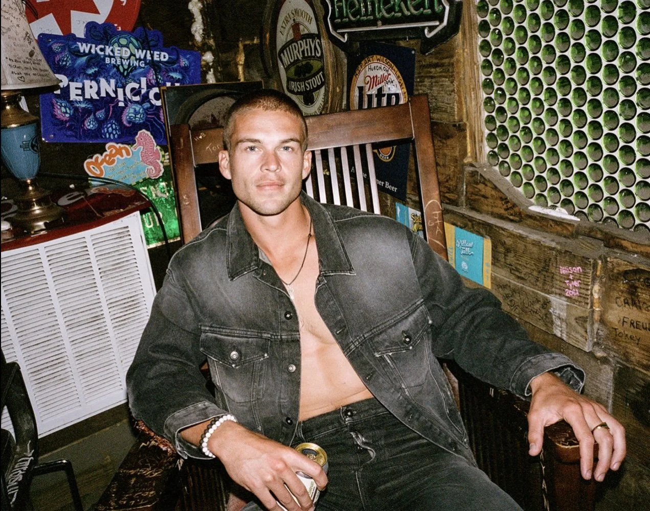 A young man sitting on a wooden chair in a bar or pub, holding a drink in his hand, wearing a black denim jacket and black pants, with a relaxed expression behind weathered wooden walls decorated with beer signs and a green circular pattern.