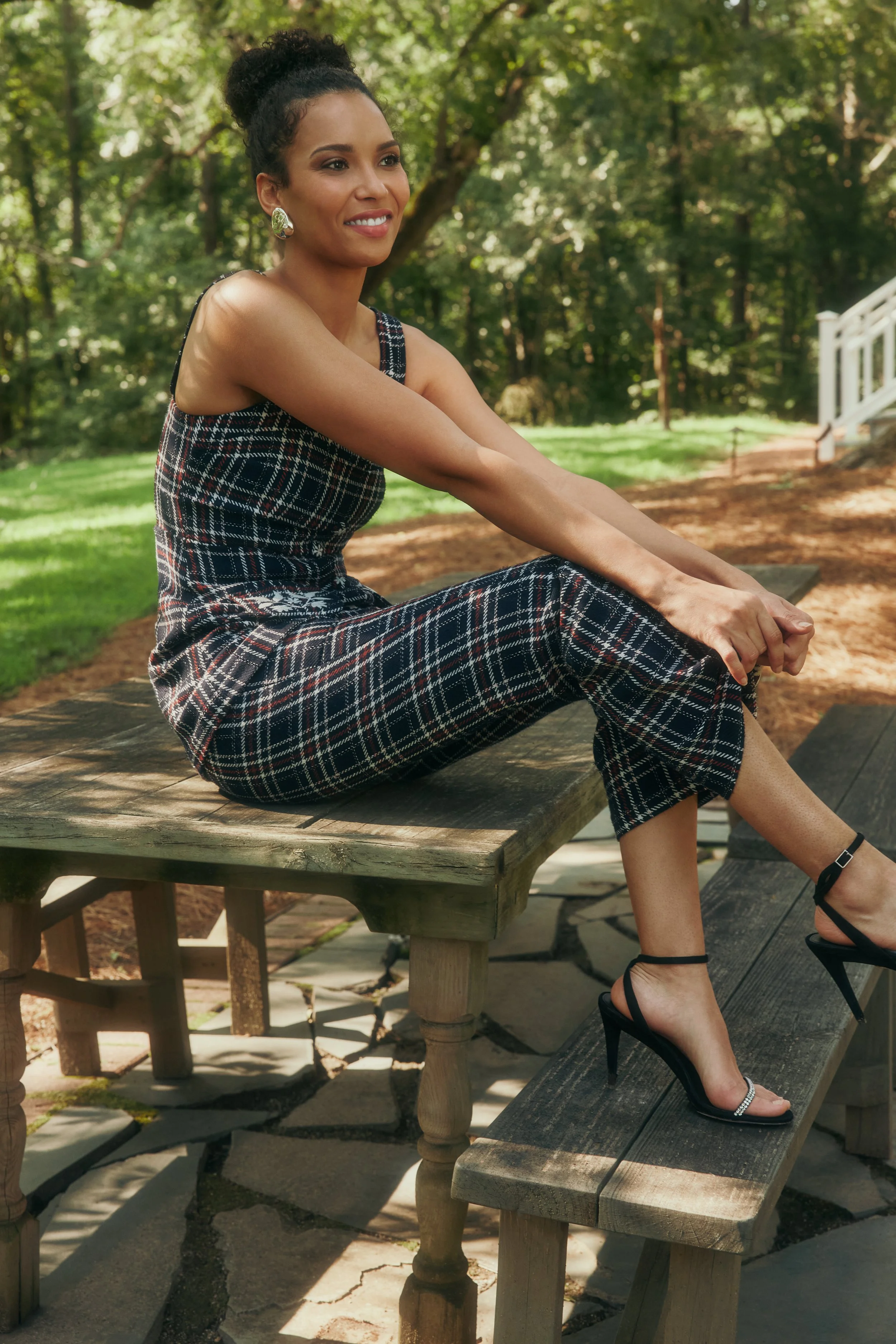 A woman sits on a wooden bench outdoors, wearing a checkered jumpsuit and black high heel sandals, with a smile, in a lush garden setting.