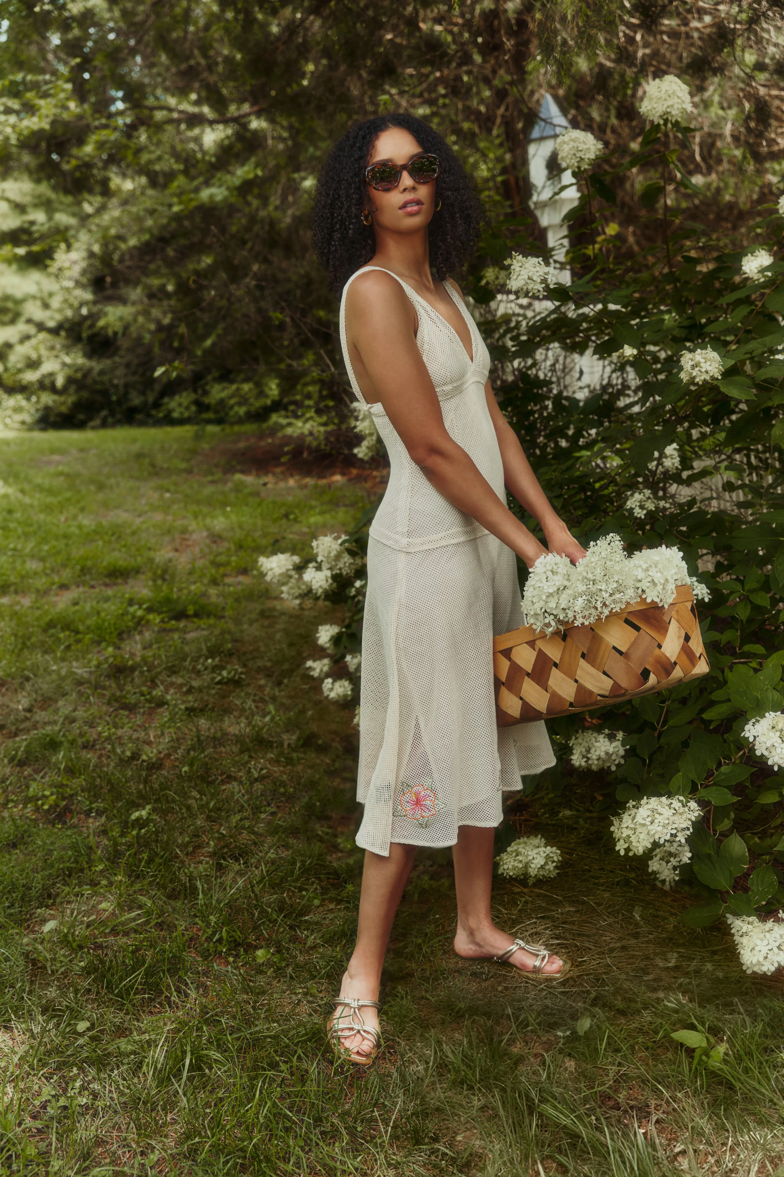 A woman wearing sunglasses and a white dress, standing outdoors in a garden with white flowers and greenery, holding a basket filled with white flowers.