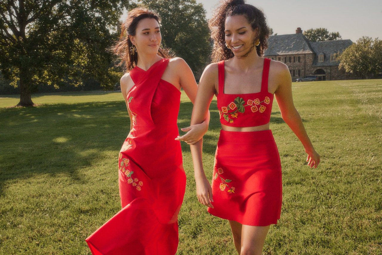 Two women in red dresses with floral embroidery walking in a grassy park area with trees and a house in the background.