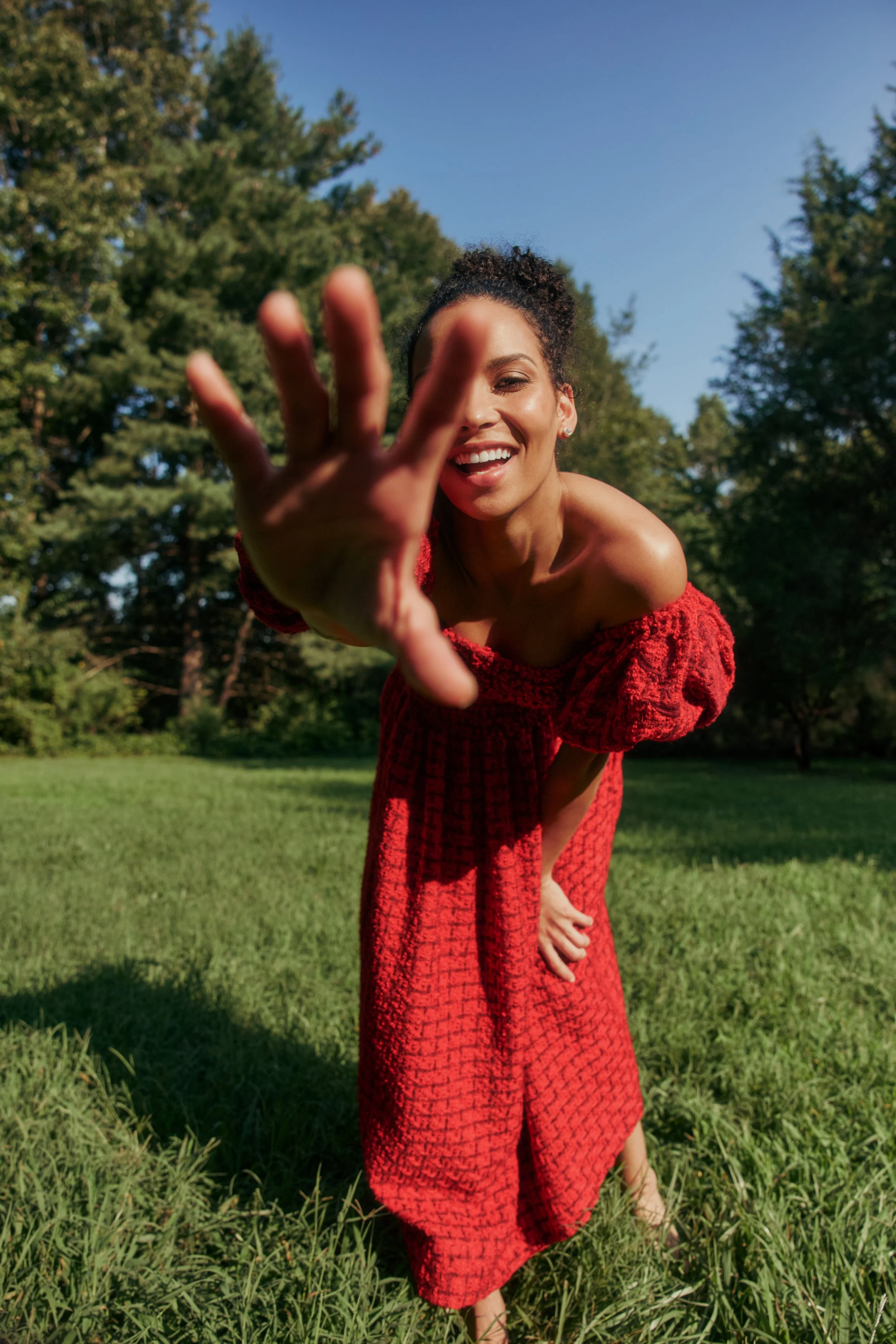 A woman in a red dress leaning slightly forward with her hand reaching towards the camera, smiling outdoors on a sunny day with green trees and grass in the background.