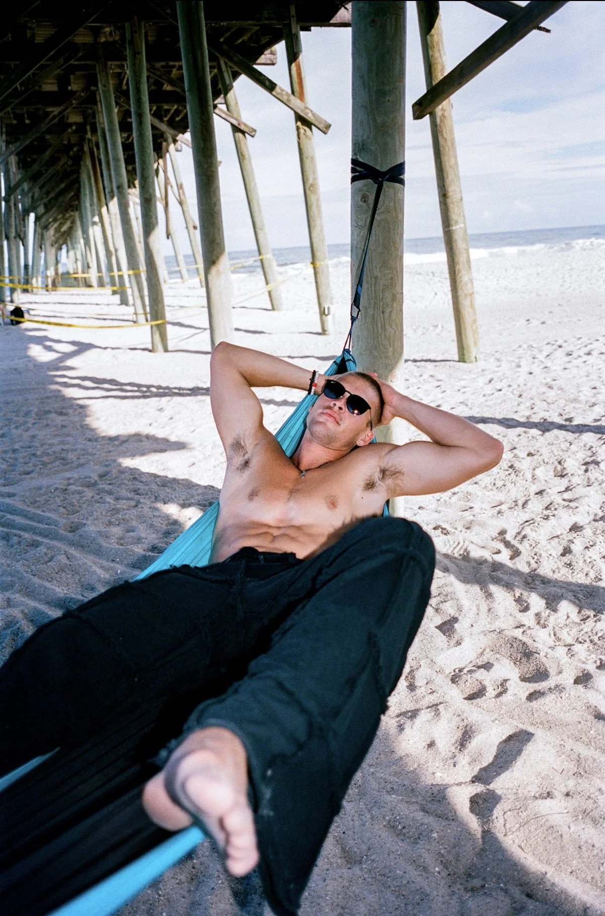 A shirtless man relaxing in a hammock under a pier on a sandy beach, wearing sunglasses and black pants, with the ocean in the background.