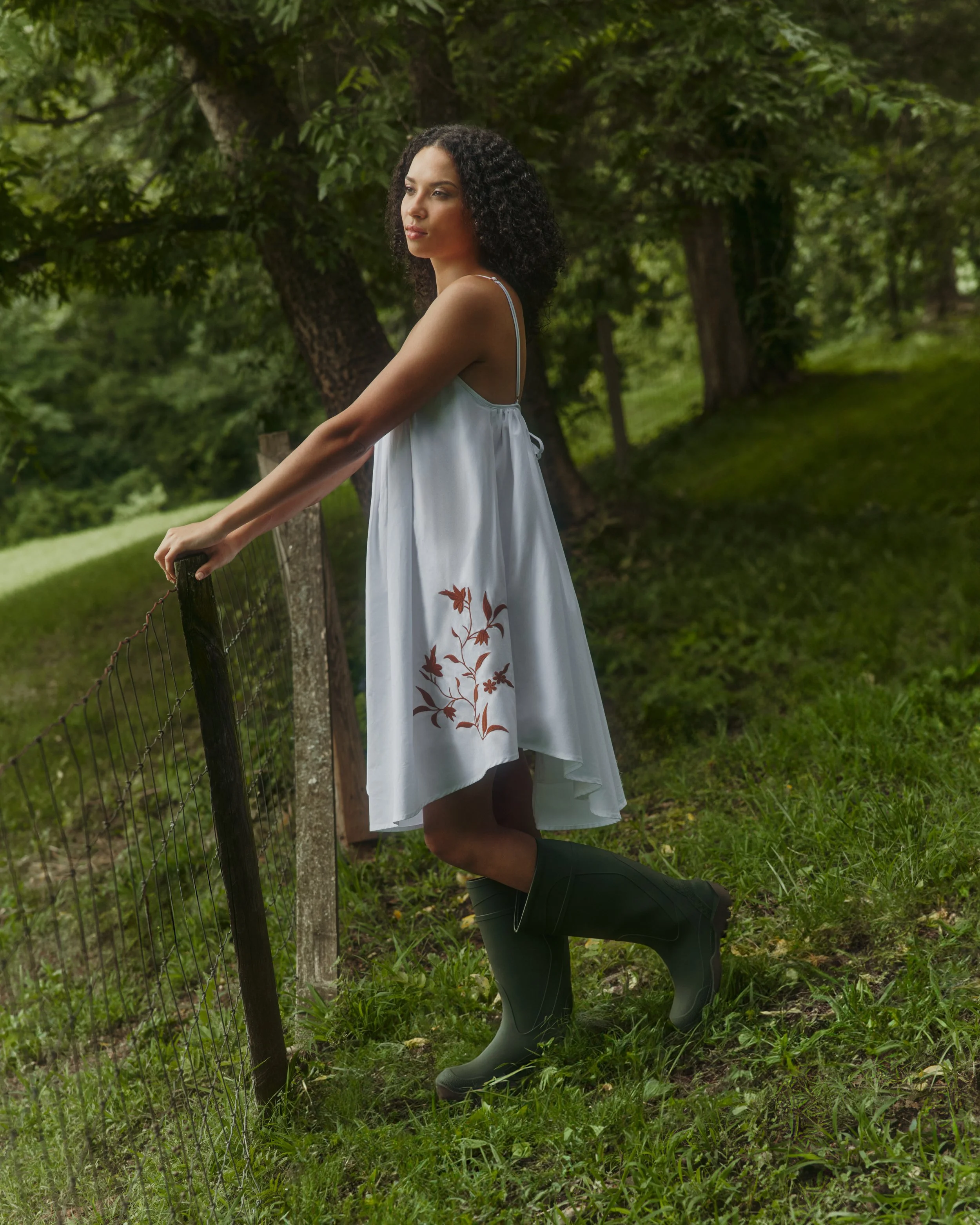 A woman wearing a white dress with brown floral embroidery, black rain boots, standing outdoors on grass, holding a fence with trees in the background.