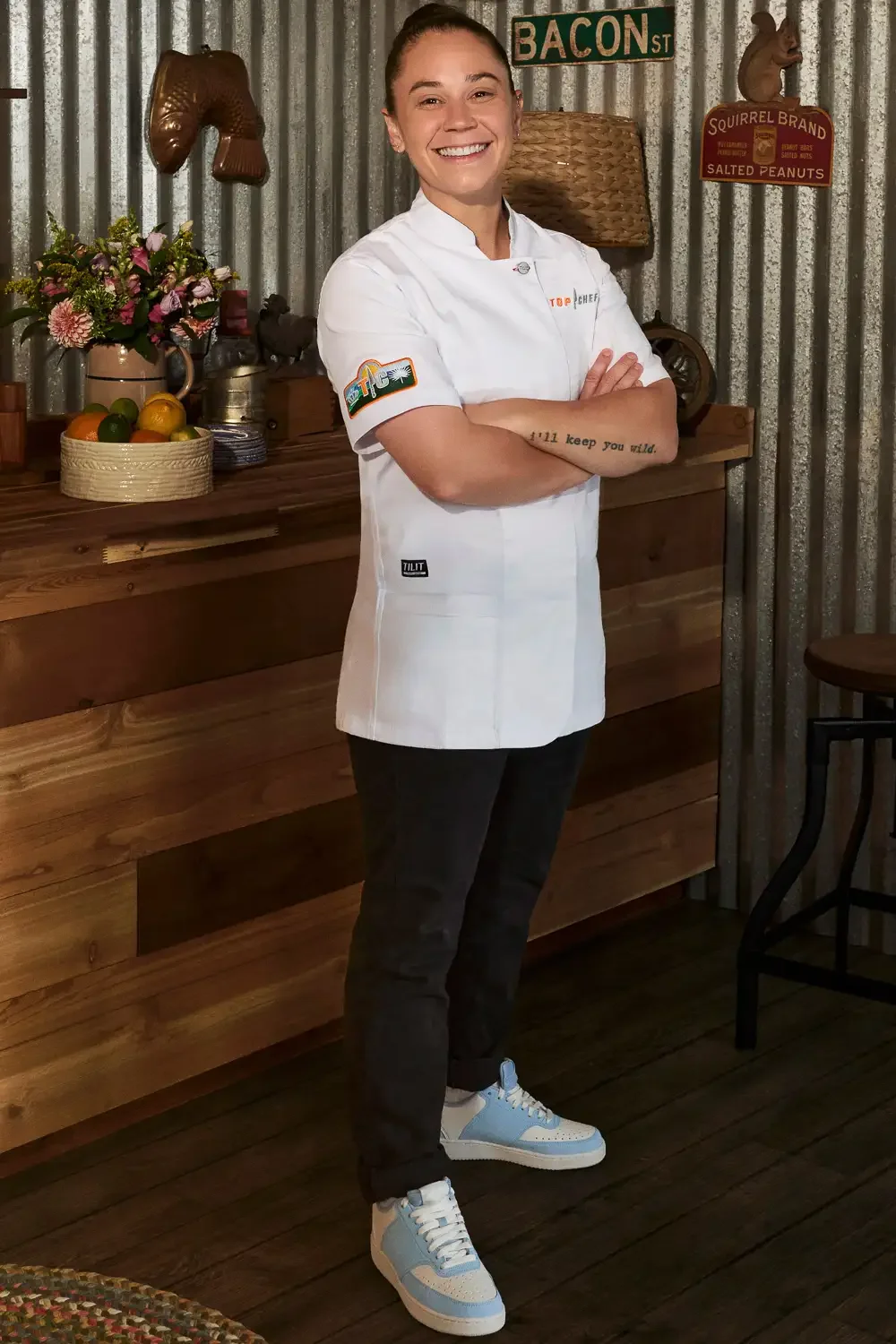 A smiling female chef wearing a white chef coat and black pants standing with arms crossed in a rustic kitchen with wooden accents and metal corrugated walls.