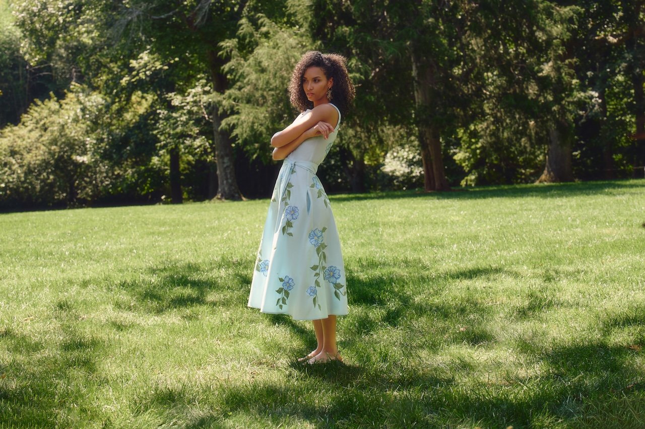 A woman standing on grass in a park, wearing a white dress with blue floral patterns, with arms crossed and looking to the side, surrounded by trees and greenery.