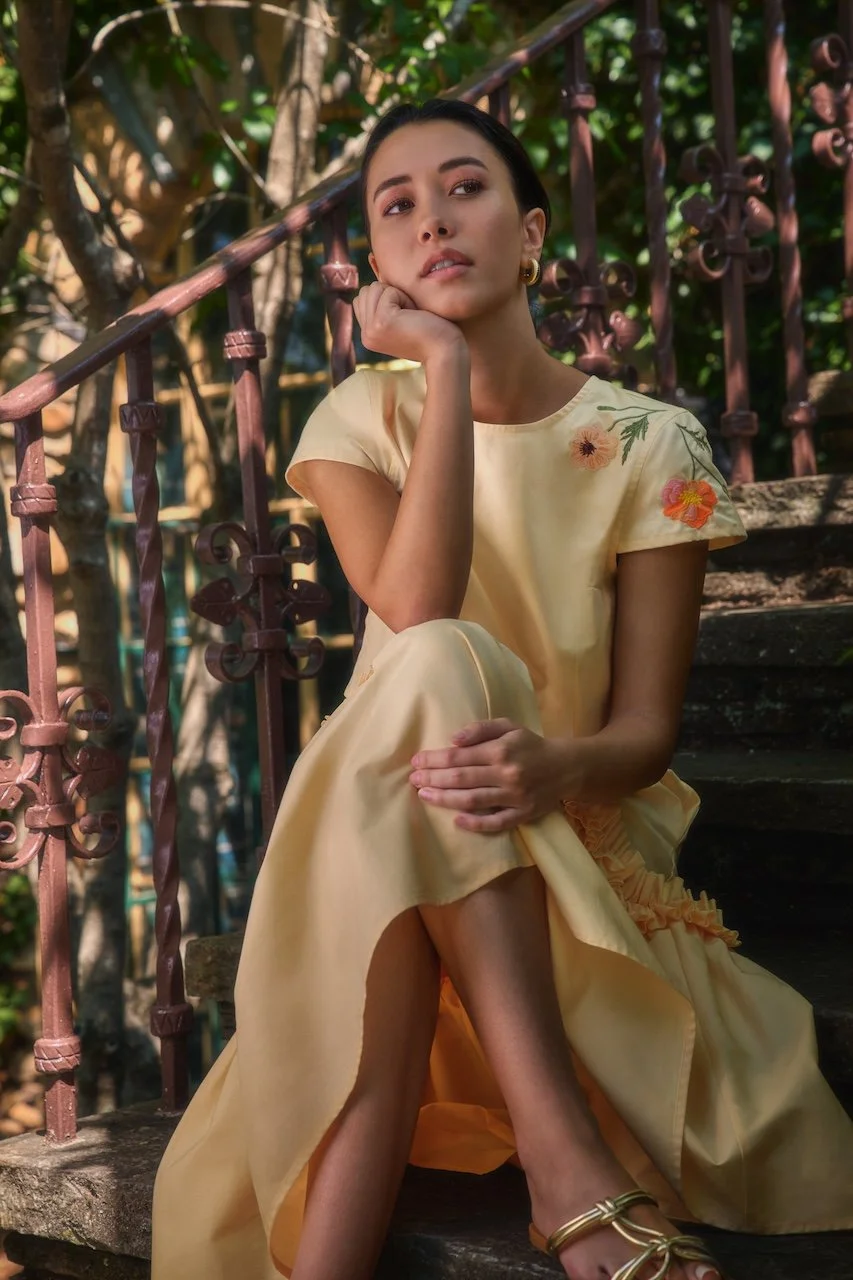 A young woman with dark hair, dressed in a pastel yellow dress with floral embroidery, is sitting on outdoor stairs with a rustic railing. She has her chin resting on her hand and is gazing thoughtfully into the distance, surrounded by greenery.