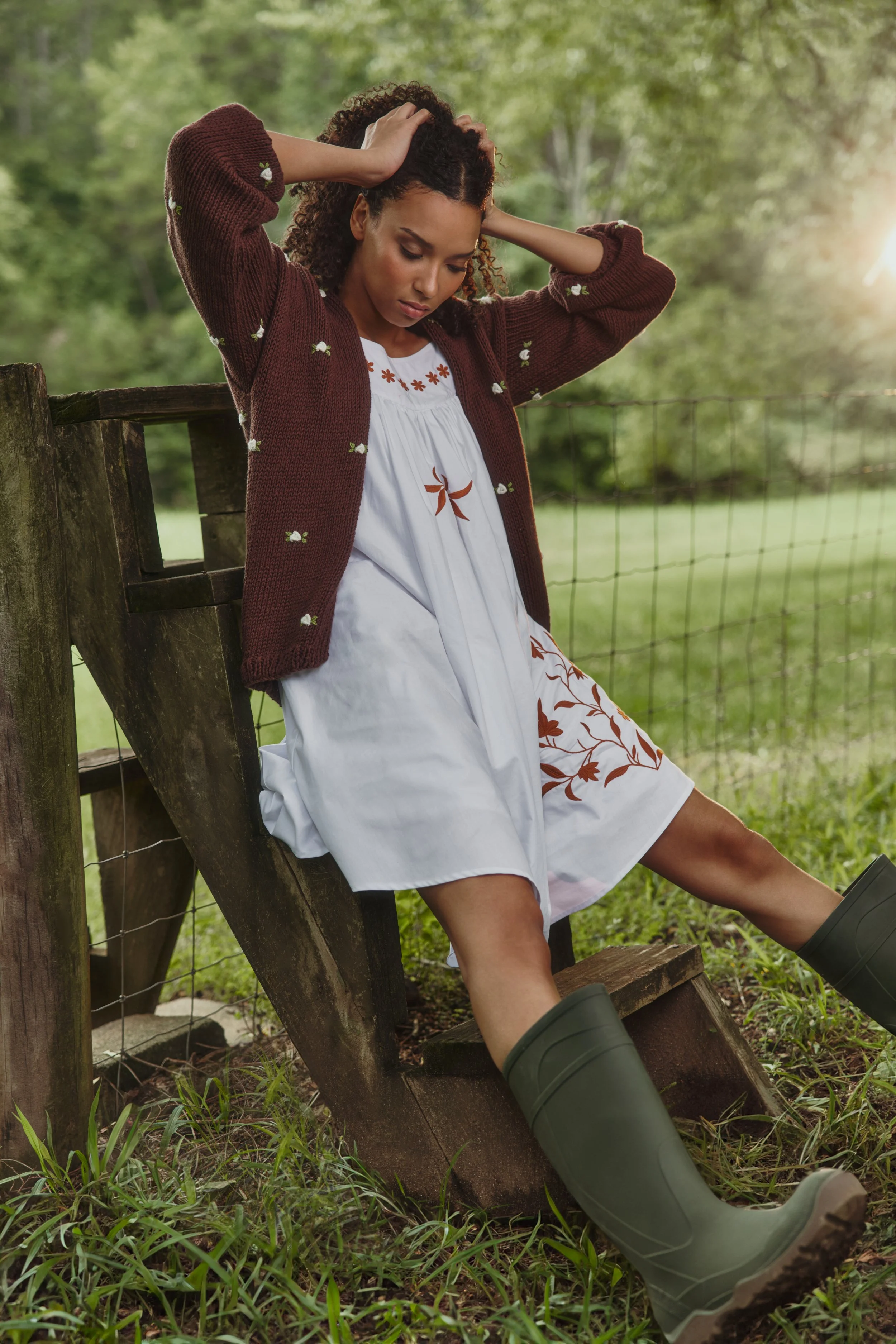 A woman sitting on a wooden fence in a grassy field, wearing a white dress with orange embroidery, a maroon cardigan, and green rubber boots, with her hands on her head and eyes closed.