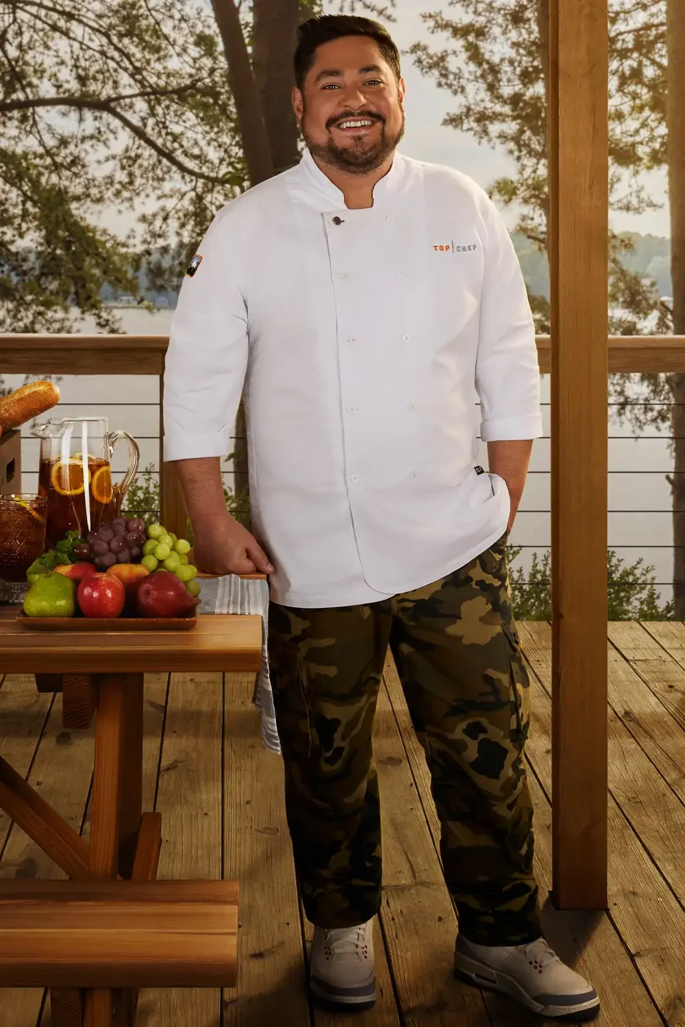 A male chef in a white coat with camouflage pants, standing outdoors on a wooden deck with trees in the background, smiling at the camera. There is a small table with fruits and a pitcher of tea nearby.