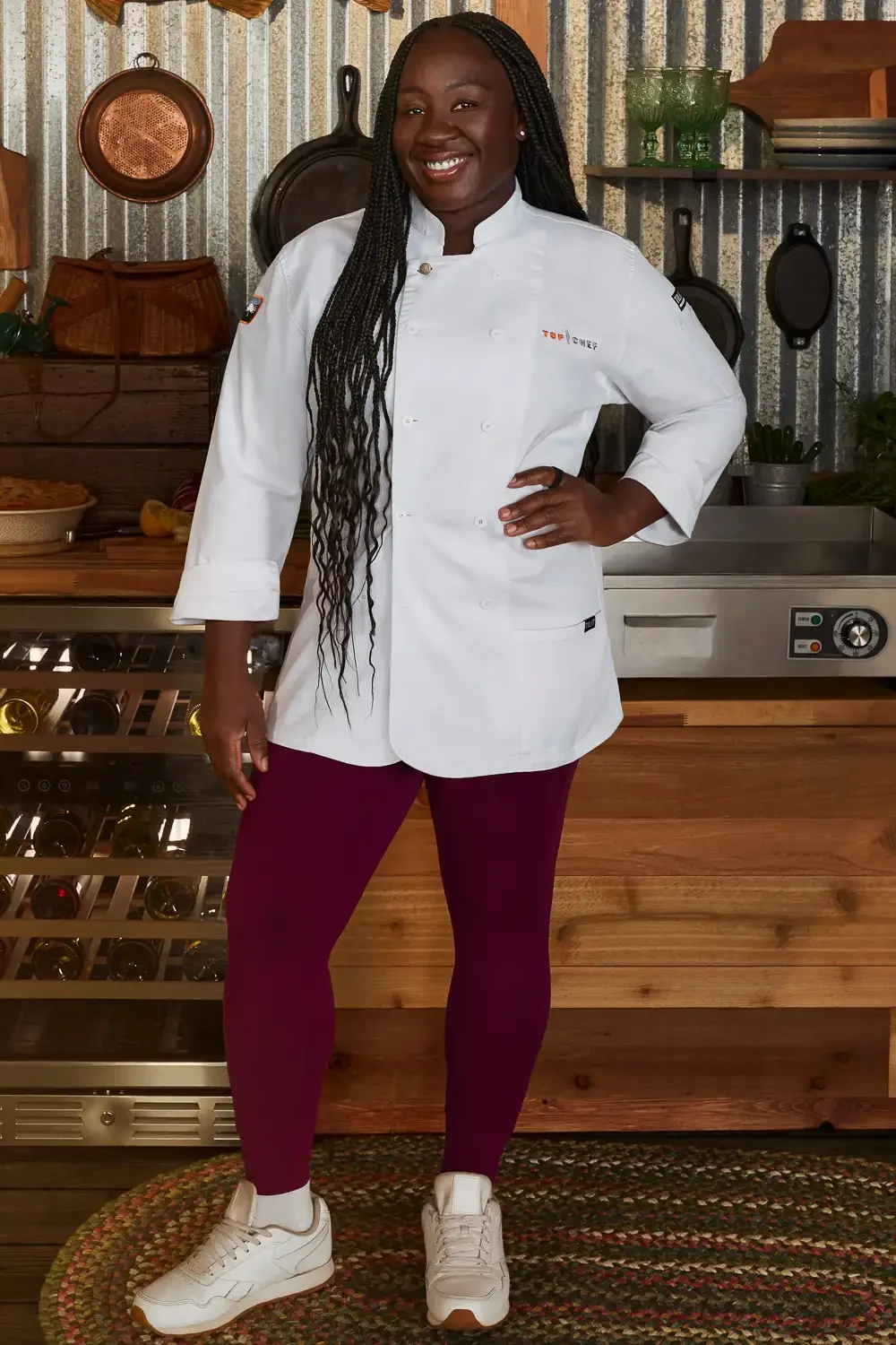 A woman in a white chef's coat, maroon leggings, and white sneakers standing in a kitchen with a rustic decor, smiling and posing with one hand on her hip.