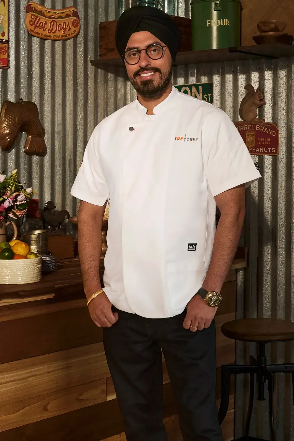 A man dressed in a white chef's coat and black turban standing in a rustic kitchen with vintage decor, including signs and wooden accents.
