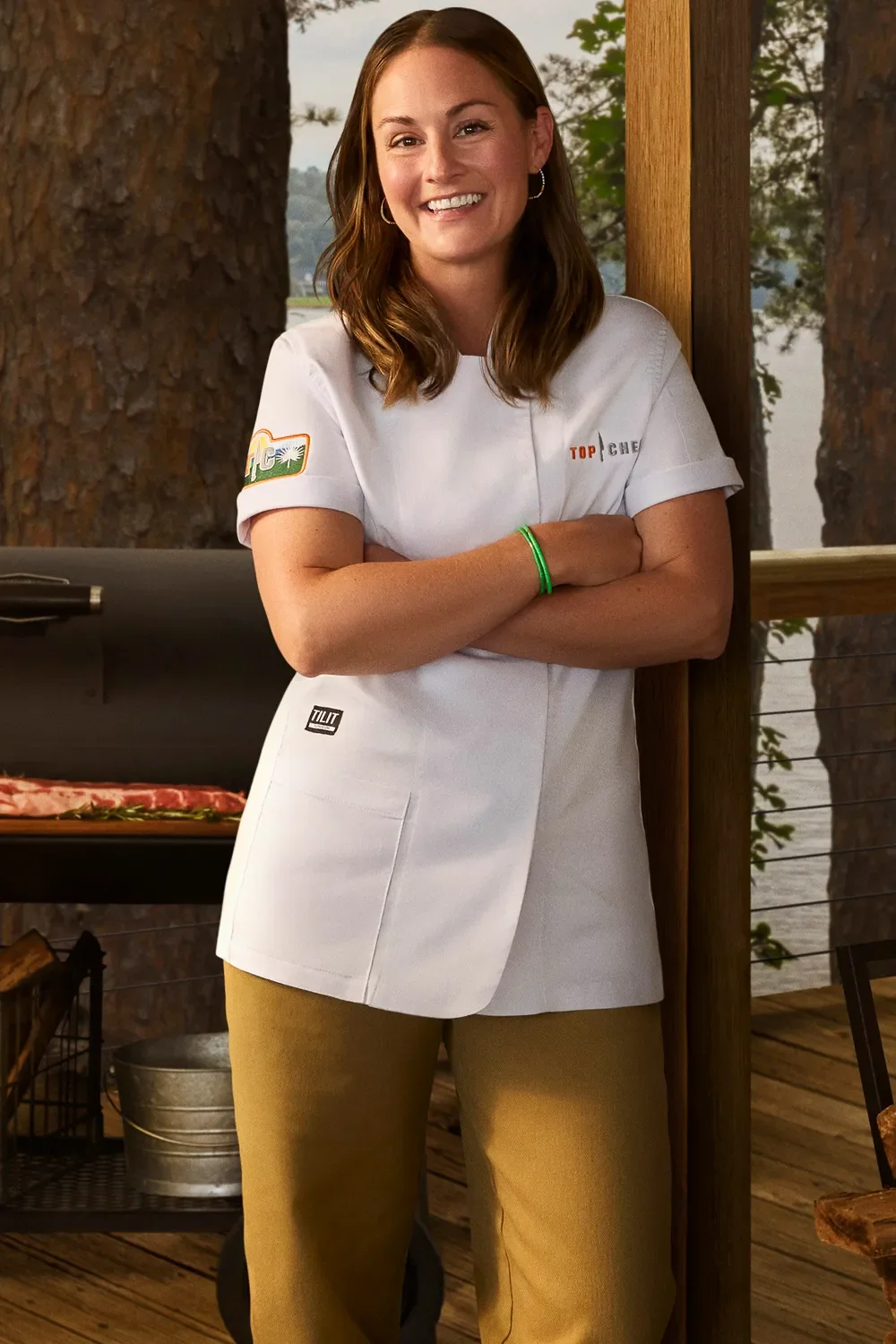A woman in a chef's white coat and tan pants, smiling with her arms crossed, standing outdoors near trees and water.