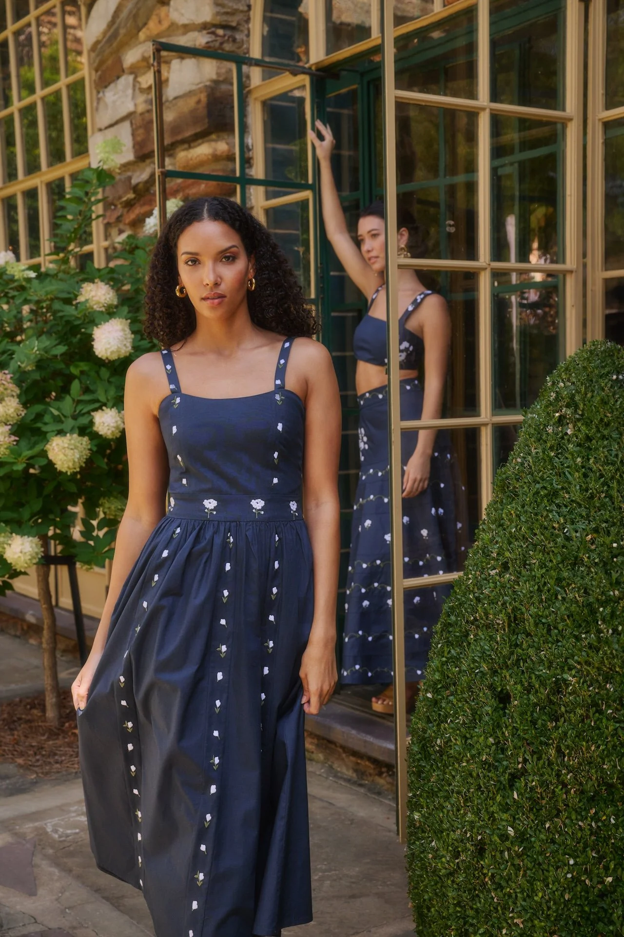 Two women standing outside a building with a large window and garden. One woman in the foreground wears a navy blue dress with white floral embroidery, and the other woman, reflected in the window, wears a similar dress.