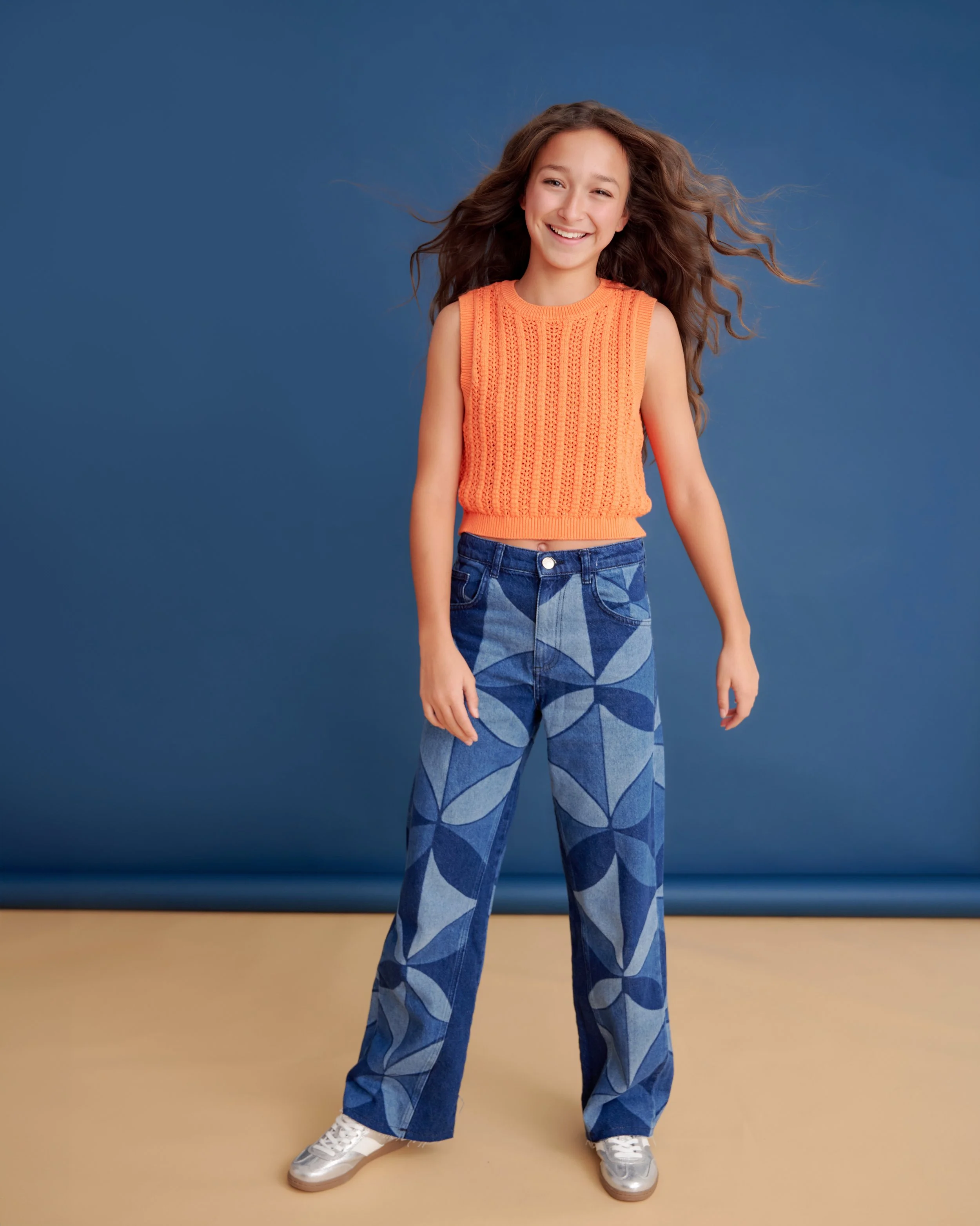 A young girl with wavy brown hair, smiling, wearing an orange sleeveless knitted top, wide-leg patterned jeans, and white sneakers, standing against a blue background.