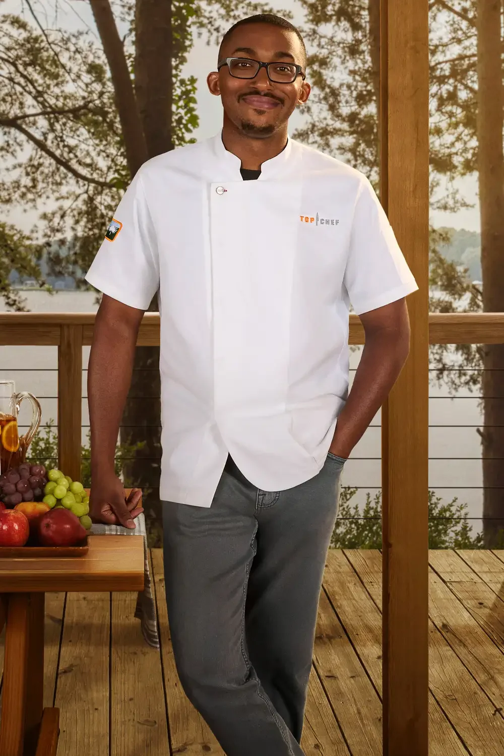 A man wearing a white chef's jacket and glasses standing on a wooden deck with trees and a lake in the background.