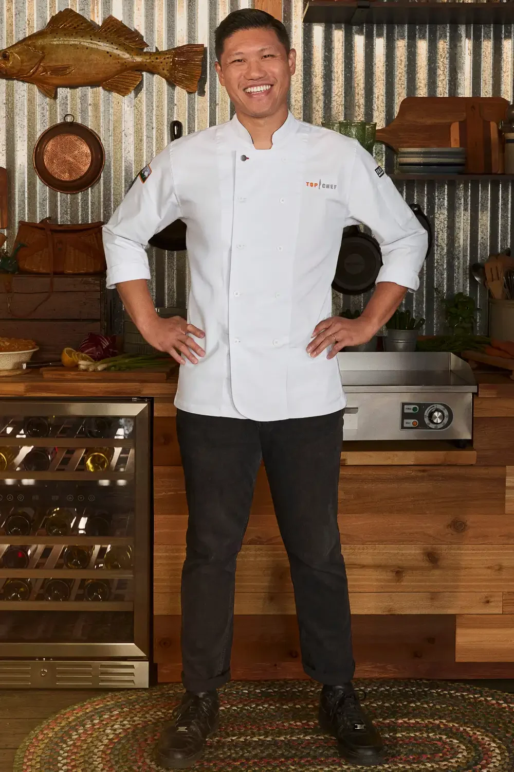A smiling male chef in a white chef's coat standing in a rustic kitchen with a fish mounted on the wall and bottles stored in a wine fridge.