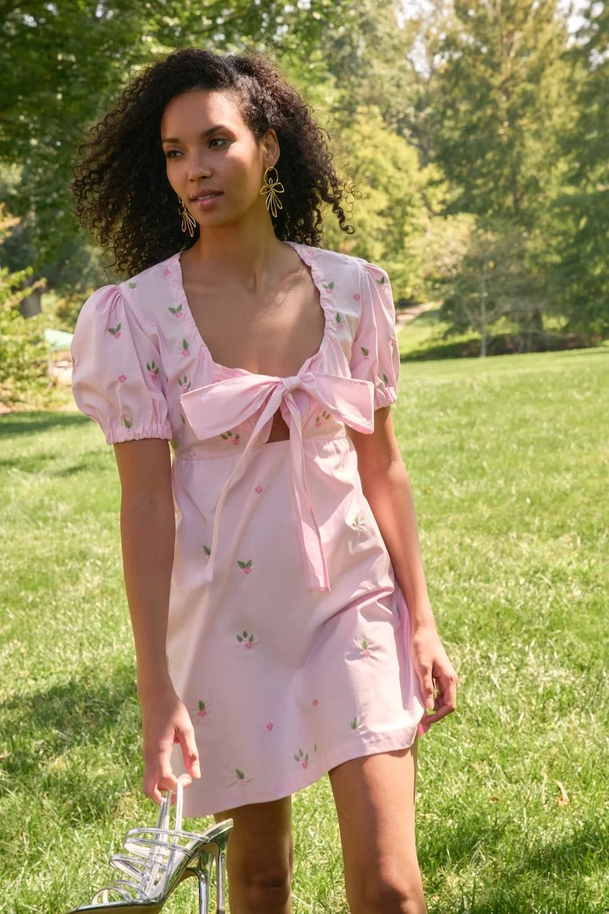 Woman with curly hair wearing a pink dress with small floral embroidery and a large bow, standing outdoors on grass with trees in the background, holding a pair of high-heeled sandals.