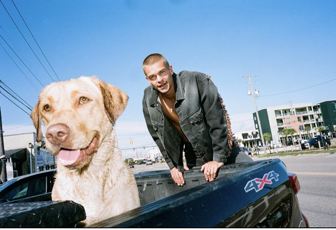 A man and a dog in the back of a pickup truck on a city street under clear blue sky.