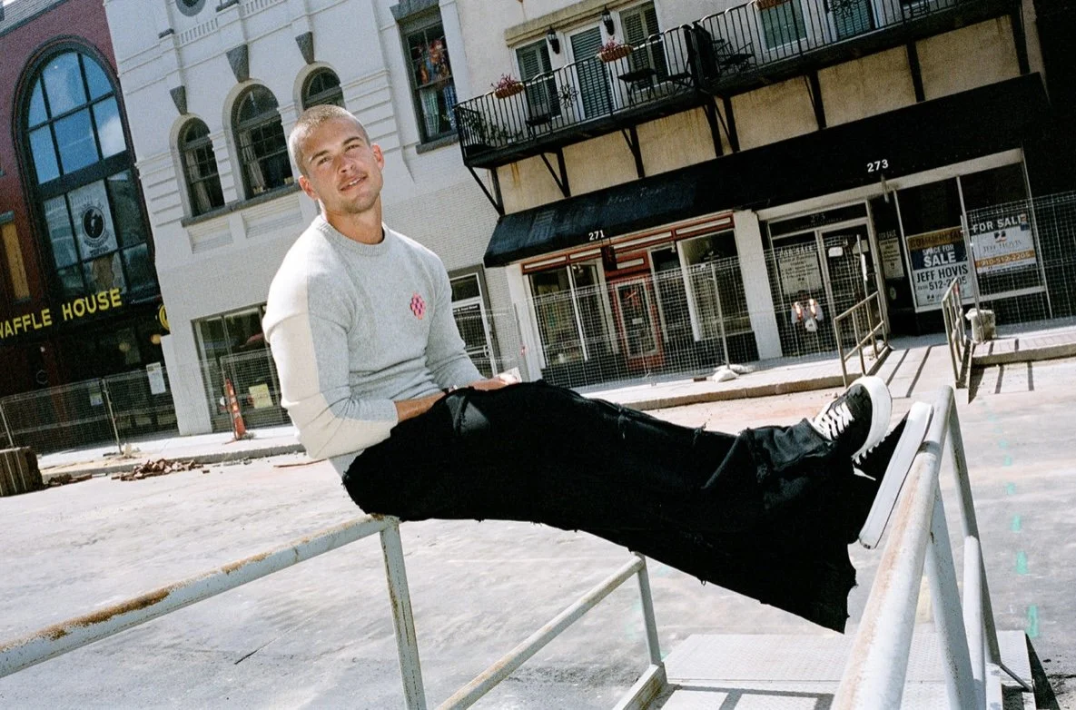A young man in a light gray sweater and black pants is sitting on a metal railing outside in a city street. There are buildings with large windows and storefronts behind him, including a waffle house and a storefront with a sale sign.