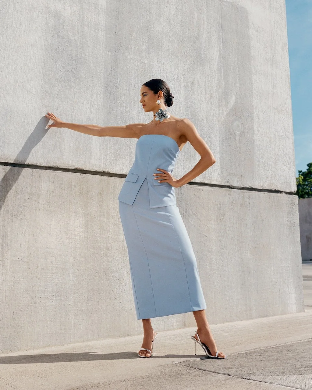 Fashionably dressed woman in a strapless blue dress and high heels stands outdoors against a beige concrete wall, with her left arm extended and touching the wall, and her right hand on her waist, posing with her gaze downward.