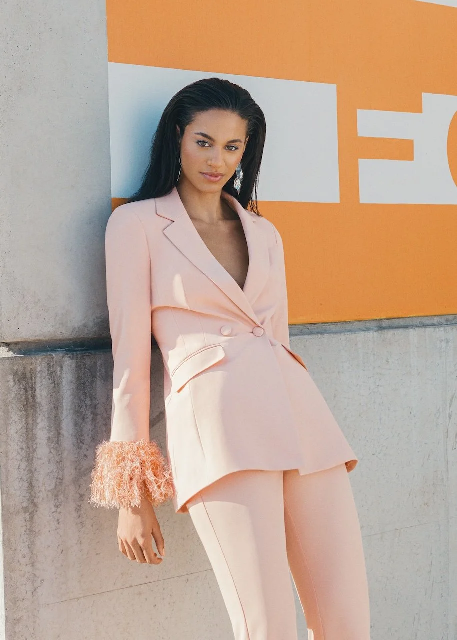 A woman in a light pink blazer and matching pants standing against a concrete wall with an orange and white sign.
