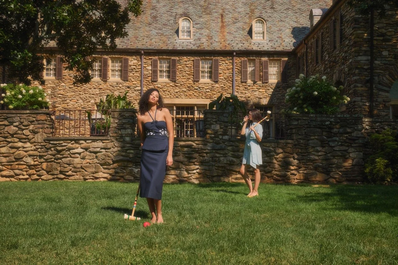 Two women playing croquet on a grassy yard in front of a stone house with multiple windows and shutters, with trees and bushes around.