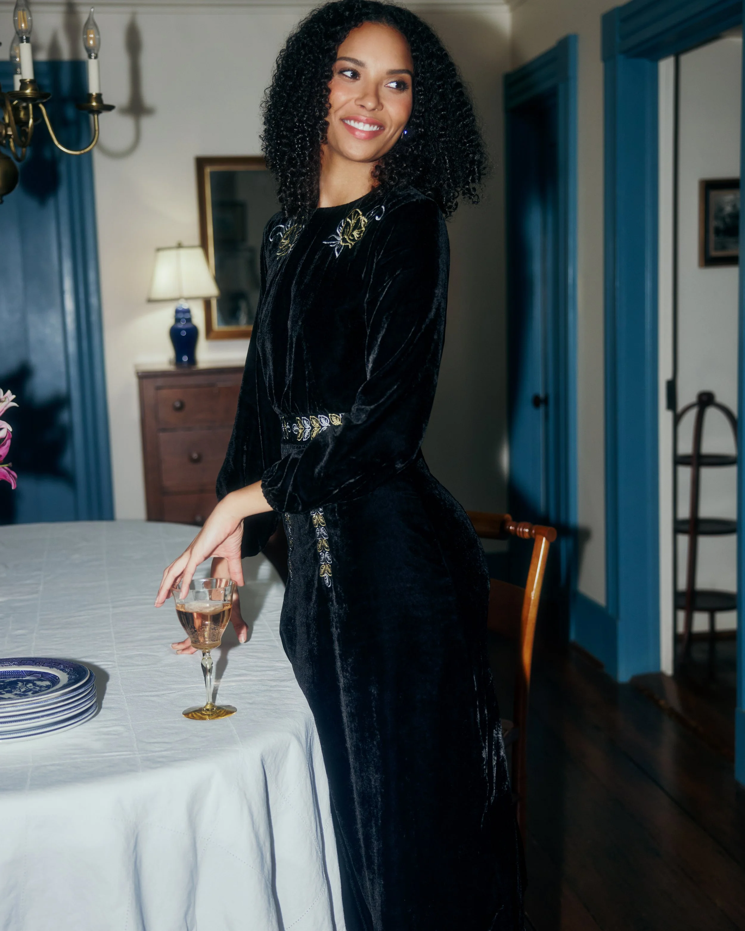 A woman with curly black hair wearing a black velvet dress with gold embroidery, holding a glass of champagne in a dining room.