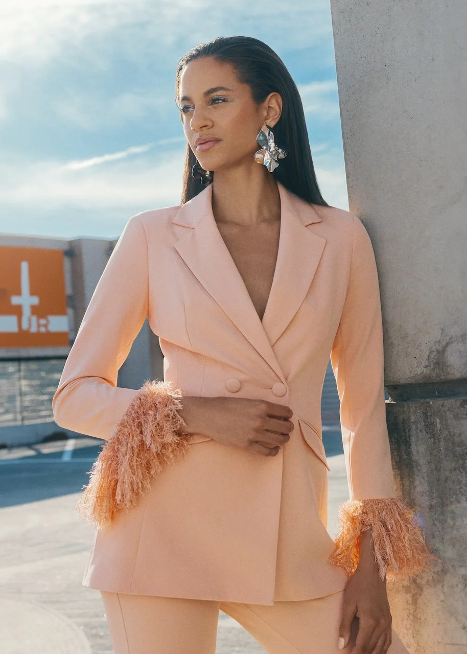 A woman with long dark hair stands outdoors against a concrete pillar, dressed in a peach blazer with feathered cuffs and matching pants, wearing large metallic earrings, with a city scene and blue sky in the background.